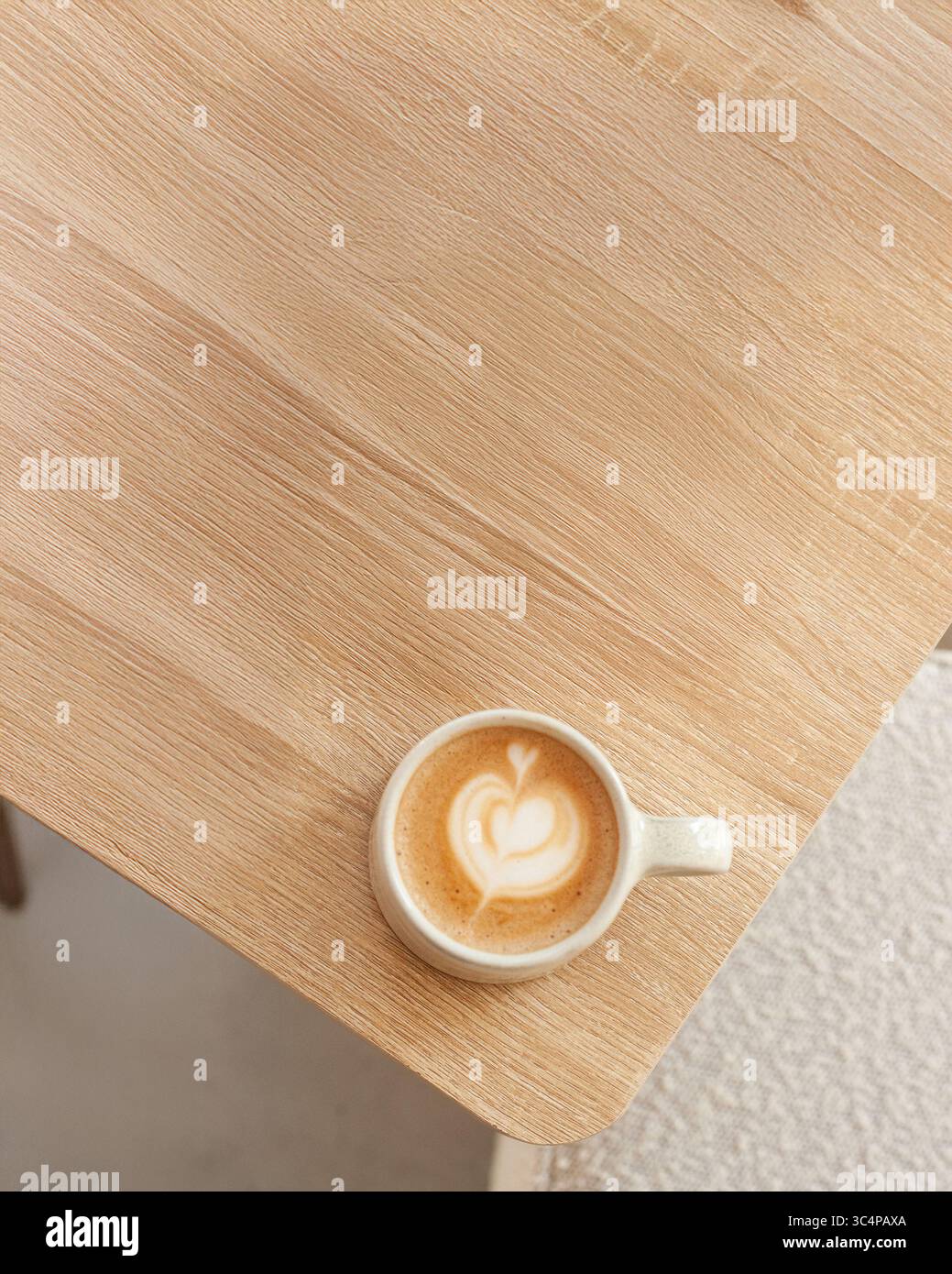 Eine Tasse Cappuccino aus Keramik mit eleganter Creme Latte Art steht auf einem hellen Holztisch in einem minimalistischen Café. Die sanfte Beleuchtung und sauber Stockfoto