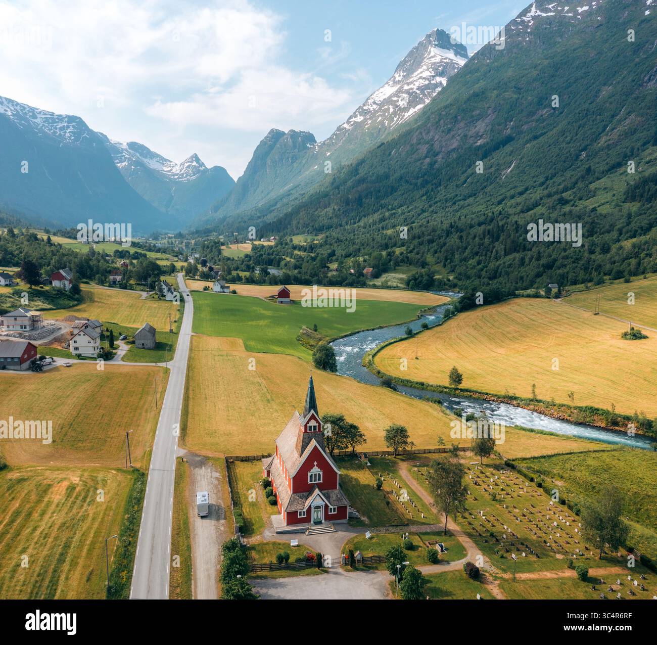 Aus der Vogelperspektive auf eine malerische rote Kirche, die stolz inmitten goldener Felder steht, mit einem sich windenden Fluss und majestätischen Bergen im Hintergrund, Olden, Vestland, Norwegen. Stockfoto