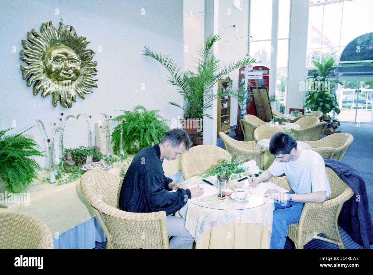 Register Entlassene Leute, Commodore, 27-08-1997 Whizgle News, Dutch Desk, Niederlande, 1950-2000 zwei Personen sitzen an einem runden Tisch in einem hellen Innenbereich und konzentrieren sich auf ihre Arbeit. Sie sind von üppigem Grün und dekorativen Elementen umgeben, darunter ein Sonnenmotiv an der Wand. Die Atmosphäre ist entspannt, mit bequemen Stühlen und natürlichem Licht. Stockfoto