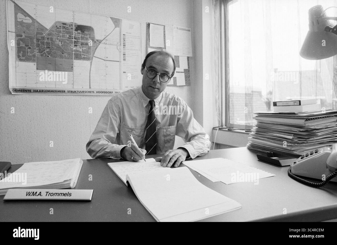 Herr TROMMEL, DORO Hoofddorp [Abteilung für Forschung und Raumentwicklung], Personen, Hoofddorp, Niederlande, 31-08-1988, Whizgle News, Dutch Desk, Niederlande, 1950-2000 Ein Mann in formeller Kleidung sitzt an einem Schreibtisch und konzentriert sich auf das Schreiben in einem Notizbuch. Hinter ihm ist eine Karte an die Wand geheftet und Stapel von Dokumenten sind ordentlich auf dem Tisch angeordnet. Eine Lampe und ein Namensschild, das ihn als WM.A. Trommels identifiziert, vervollständigen die Szene. Stockfoto