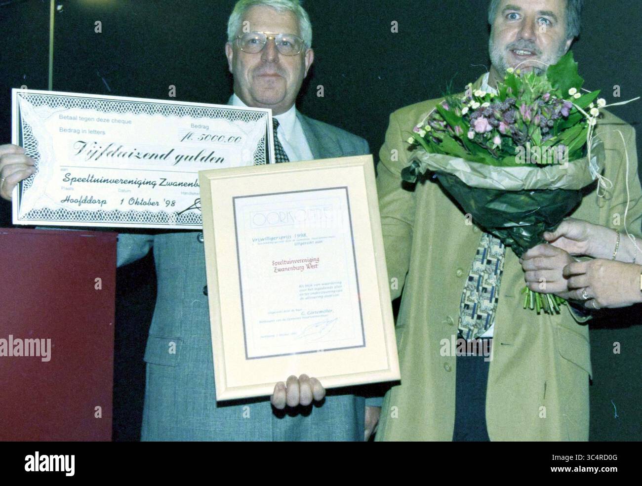 Volunteer Award, Haarlemmermeer, 30-09-1998, Whizgle News, Dutch Desk, Niederlande, 1950-2000 zwei Männer stehen zusammen, einer mit einem großen Scheck, der andere mit einem gerahmten Zertifikat und einem Blumenstrauß. Sie scheinen eine Leistung zu feiern, möglicherweise eine Auszeichnung oder Anerkennung für ein Unternehmen oder eine Einzelperson. Der Mann mit dem Scheck lächelt weit, während der andere zufrieden und engagiert aussieht. Stockfoto