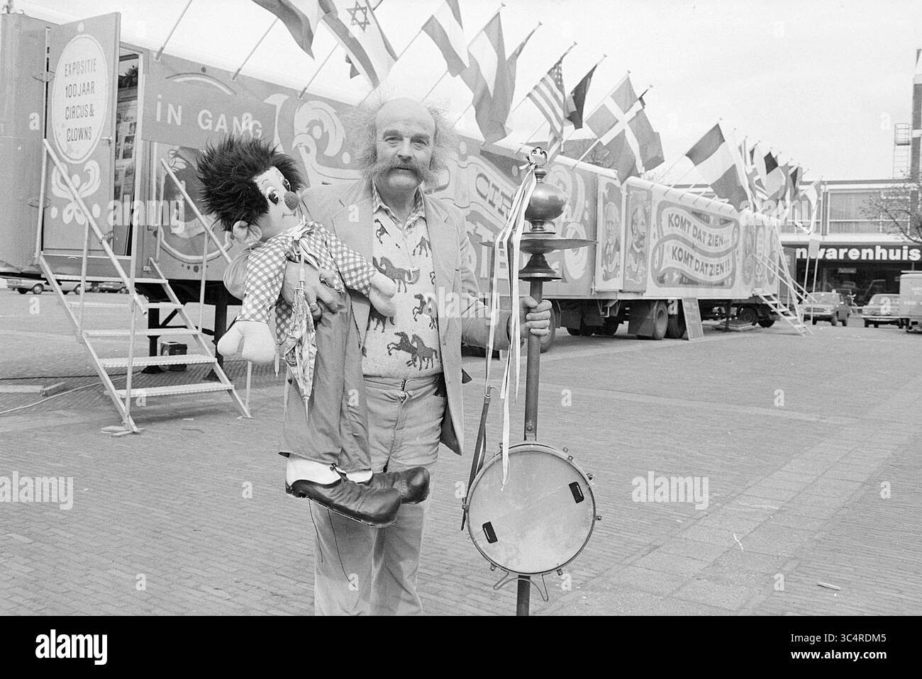 Johnny (Boltini) Akkerman für Zirkusmuseum, Circus, 17-04-1977 Whizgle News, Dutch Desk, Niederlande, 1950-2000 Ein Mann mit markantem Schnurrbart hält eine Puppe in der einen Hand und ein Banjo in der anderen, vor bunten Fahnen und einem dekorierten Truck in einer lebhaften Umgebung im Freien. Stockfoto