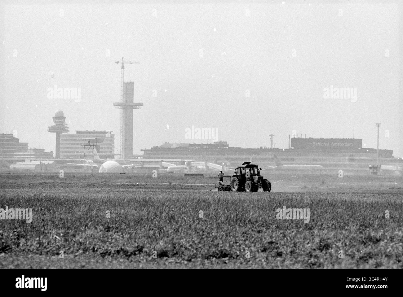 Verschiedene rund um Schiphol, Schiphol, Flugzeuge, Schiphol, 25-05-1989 Whizgle News, Dutch Desk, Niederlande, 1950-2000 Ein Traktor befährt die Felder im Vordergrund, während im Hintergrund ein Flughafen mit hohen Terminalgebäuden und Kontrolltürmen den Kontrast zwischen Landwirtschaft und modernem Verkehr unterstreicht. Stockfoto
