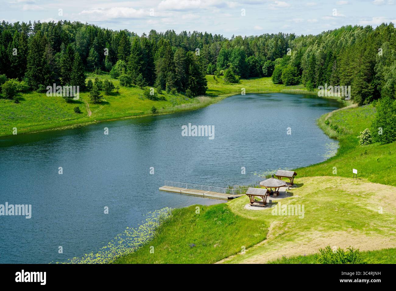 Ein ruhiger Blick auf den See in der Nähe von Stanczyki, Polen, mit hölzernen Picknickunterkünften, einem Dock, üppigem Grün und bewaldeten Hügeln unter einem sonnigen Himmel Stockfoto