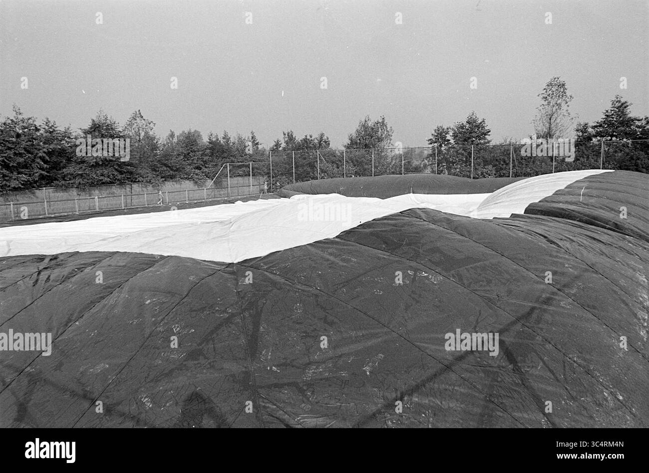 Aufblasbare Sporthalle Tennisplatz, 00-10-1969 Whizgle News, Dutch Desk, Niederlande, 1950-2000 A groß, die aufgeblasene Stoffstruktur liegt auf dem Boden auf und zeigt eine geschwungene und mehrlagige Oberfläche. In der Umgebung gibt es Bäume und einen schwachen Umriss eines Sportplatzes im Hintergrund, was eine ruhige Outdoor-Szene schafft. Stockfoto