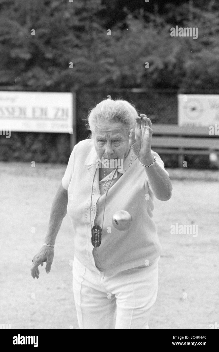 Frau Steenman jeu de Boule in Boulodrome, Jeu de Boule, 19-08-1987 Whizgle News, Dutch Desk, Niederlande, 1950-2000 eine ältere Frau mit entschlossenem Ausdruck ist in der Mitte der Bewegung, wirft fachmännisch eine Metallkugel auf ein Ziel auf den Boden und zeigt ihr Können in einem Pétanque-Spiel. Sie trägt ein leichtes Oberteil und eine Stoppuhr um den Hals, was ihre Rolle im Spiel anzeigt. Bäume und Bänke bilden die Kulisse und tragen zur entspannten Atmosphäre der Szene bei. Stockfoto