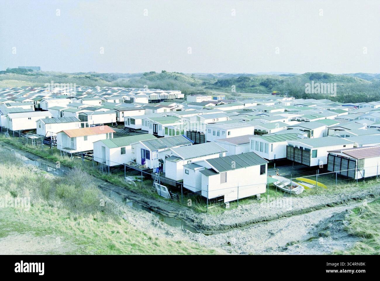 Beach Houses, IJmuiden, IJmuiden, Niederlande, 11-03-2002 Whizgle News, Dutch Desk, Niederlande, 1950-2000 Eine weitläufige Landschaft aus übersichtlich angeordneten, modularen Häusern erstreckt sich über eine natürliche Umgebung mit sanften Hügeln im Hintergrund. Das Layout spiegelt eine Gemeinschaftsatmosphäre wider und zeigt eine Mischung aus kleinen Häusern und Wohnwagen, die alle in unmittelbarer Nähe angeordnet sind. Grüne Dächer stehen im Kontrast zu den neutralen Außenflächen, während Grasflecken und Vegetationsbüschel die Monotonie der Strukturen durchbrechen. Stockfoto