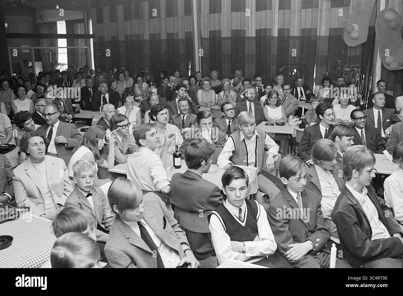 Papierhandelsschuldiplome, Präsentation, 10-06-1969 Whizgle News, Dutch Desk, Niederlande, 1950-2000 Eine große Menschenmenge versammelt sich in einem Saal, an Tischen mit karierten Tischdecken, in Gesprächen oder aufmerksamer Beobachtung der Front. Das Publikum umfasst eine Mischung aus Erwachsenen und Jugendlichen, die alle in formeller Kleidung gekleidet sind und eine besondere Veranstaltung oder Feier vorschlagen. Stockfoto