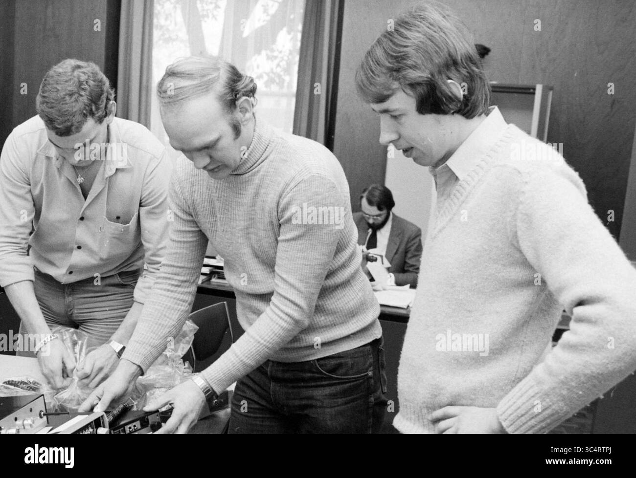 Konfiszierte Sendegeräte bei der Polizeistation Haarlem, Sender, Sendegeräte, Haarlem, Niederlande, 05-02-1980 Whizgle News, Dutch Desk, Niederlande, 1950-2000 drei Personen konzentrieren sich auf ein technisches Projekt, bei dem sie an einem auf einem Tisch liegenden Gerät zusammenarbeiten. Eine Person, die einen Pullover trägt, arbeitet am Gerät, während die beiden anderen aufmerksam beobachten. Im Hintergrund ist eine andere Person an ihrer eigenen Aufgabe beteiligt, was eine dynamische und kollaborative Umgebung veranschaulicht. Stockfoto