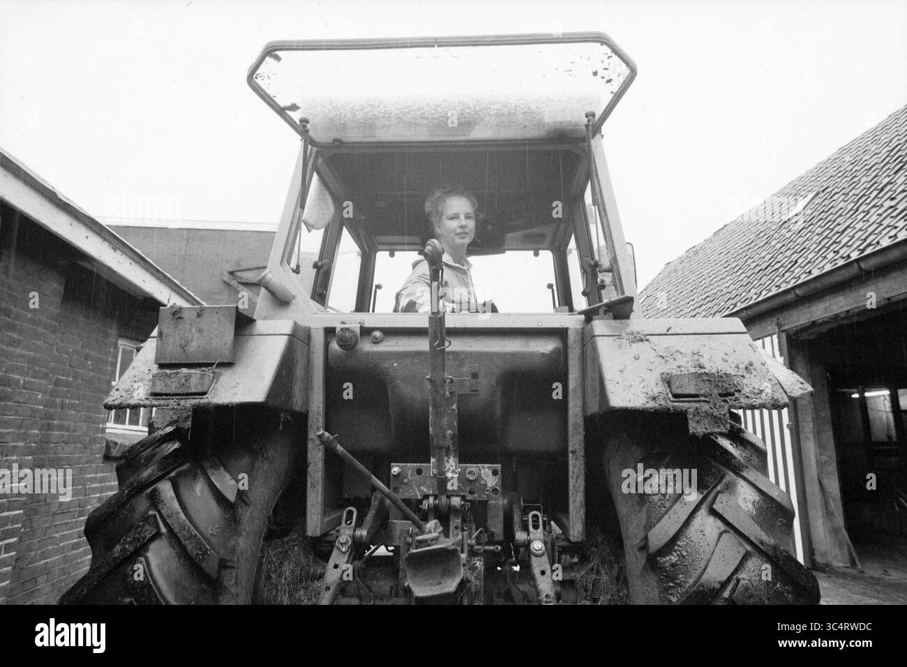 Farmer Jose van Geldrop, 29-09-1991 Whizgle News, Dutch Desk, Niederlande, 1950-2000 Eine junge Frau sitzt selbstbewusst am Lenkrad eines Traktors, umgeben von rustikalen Bauernhäusern. Die matschigen Reifen deuten auf einen Tag harter Arbeit hin, während der bewölkte Himmel der Szene einen düsteren Ton verleiht. Stockfoto