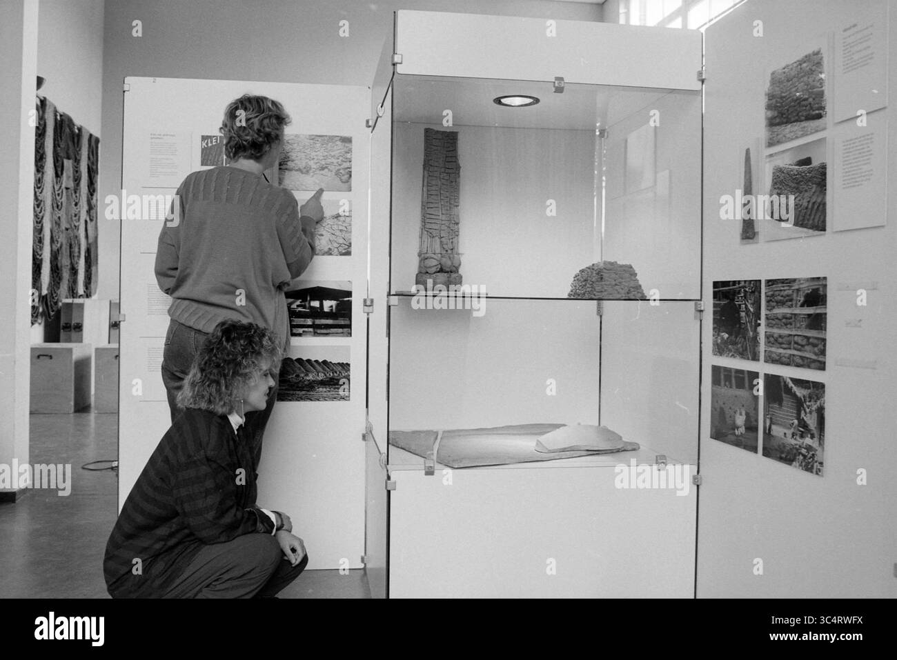 Pottery-Ausstellung, 00-00-1988 Whizgle News, Dutch Desk, Niederlande, 1950-2000 zwei Personen sind in einem Ausstellungsraum beschäftigt und untersuchen Vitrinen mit Artefakten. Eine Person zeigt auf die Informationen auf einem Brett, während die andere sich in der Hocke befindet, um die Gegenstände genau zu beobachten. Neben der Ausstellung befinden sich verschiedene Bilder und Beschreibungen an den Wänden, die zur lehrreichen Atmosphäre der Ausstellung beitragen. Stockfoto
