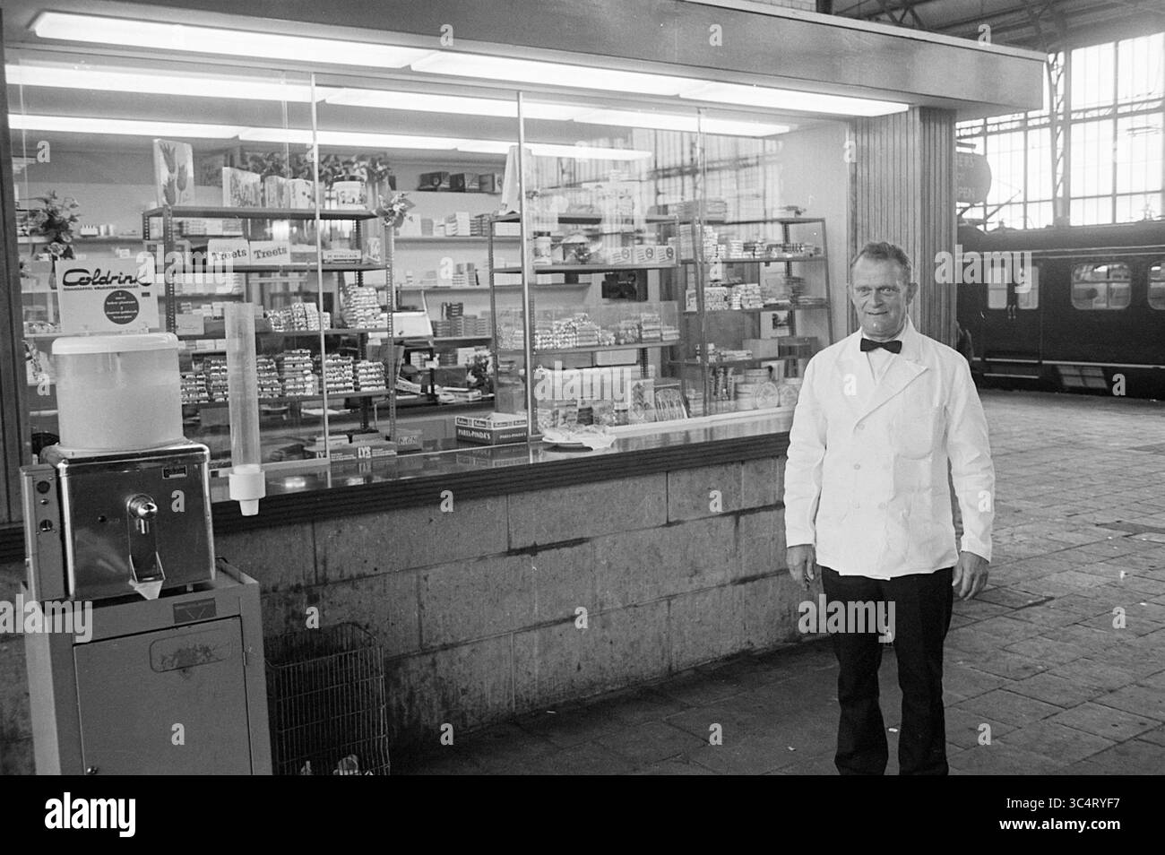 Mr. Cop am Süßwarenstand, Personen, 25-06-1968 Whizgle News, Dutch Desk, Niederlande, 1950-2000 Ein Oldtimer steht stolz vor einer gut sortierten Theke mit verschiedenen Artikeln, darunter verpackte Waren und Snacks. Hinter ihm ist ein Zug sichtbar, der auf die geschäftige Umgebung eines Bahnhofs oder Marktes hinweist. Der Ladenbesitzer trägt eine Schleife und einen knusprigen weißen Mantel, der einen klassischen, nostalgischen Charme verkörpert. Stockfoto
