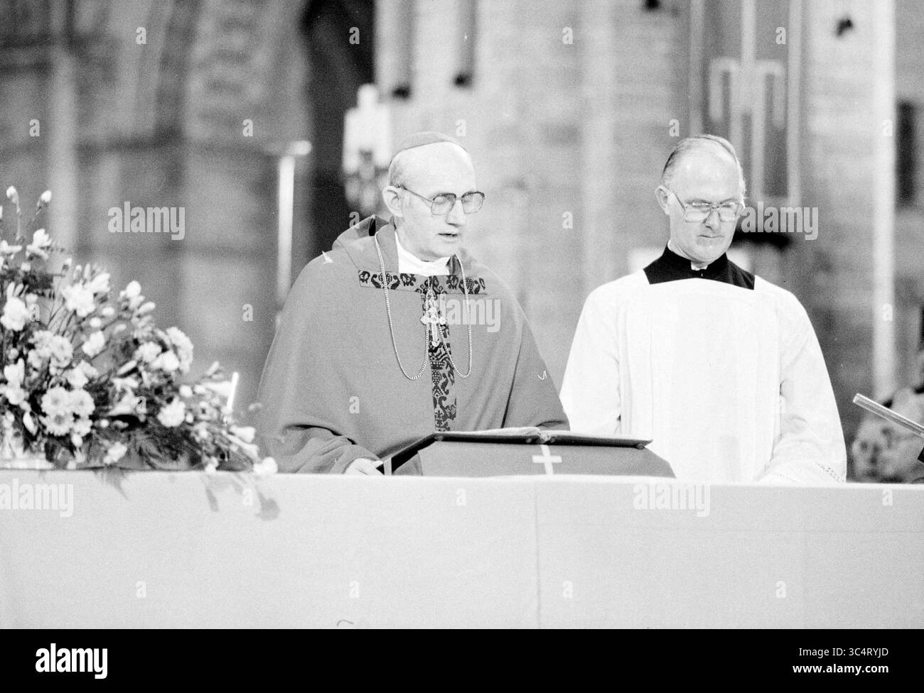 Bischof Bomers und Weihbischof Lescrauwaert, RK Bavo, Haarlem, Weihe, römisch-katholische Kirche, Haarlem, Niederlande, 14-01-1984 Whizgle News, Dutch Desk, Niederlande, 1950-2000 Ein Klerus hält eine Predigt bei einer religiösen Zeremonie, begleitet von einem anderen Kleriker. Das Ambiente ist mit Blumenarrangements und religiösen Symbolen versehen, die eine feierliche Atmosphäre schaffen. Stockfoto