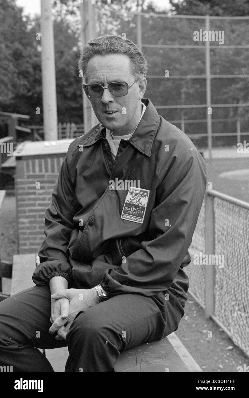 Softball, Dries de Zwaan, Softball, 07-10-1978 Whizgle News, Dutch Desk, Niederlande, 1950-2000 Ein Mann sitzt in Sportkleidung auf einer Bank, trägt eine Sonnenbrille und eine Jacke mit einem Abzeichen. Er hat ordentlich Haare gekämmt und wirkt besinnlich, mit einem Baseballfeld und Fechten im Hintergrund. Stockfoto