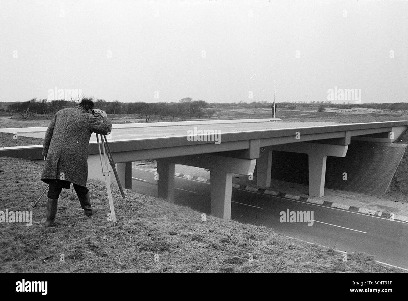 Bauviadukt, Noordwijkerhout, Baugruben, 05-03-1970 Whizgle News, Dutch Desk, Niederlande, 1950-2000 Eine Person verwendet ein Vermessungsinstrument auf einem Stativ, um die Struktur einer Brücke zu messen und zu bewerten, die eine ruhige Straße überspannt und von einer kargen, offenen Landschaft umgeben ist. Stockfoto