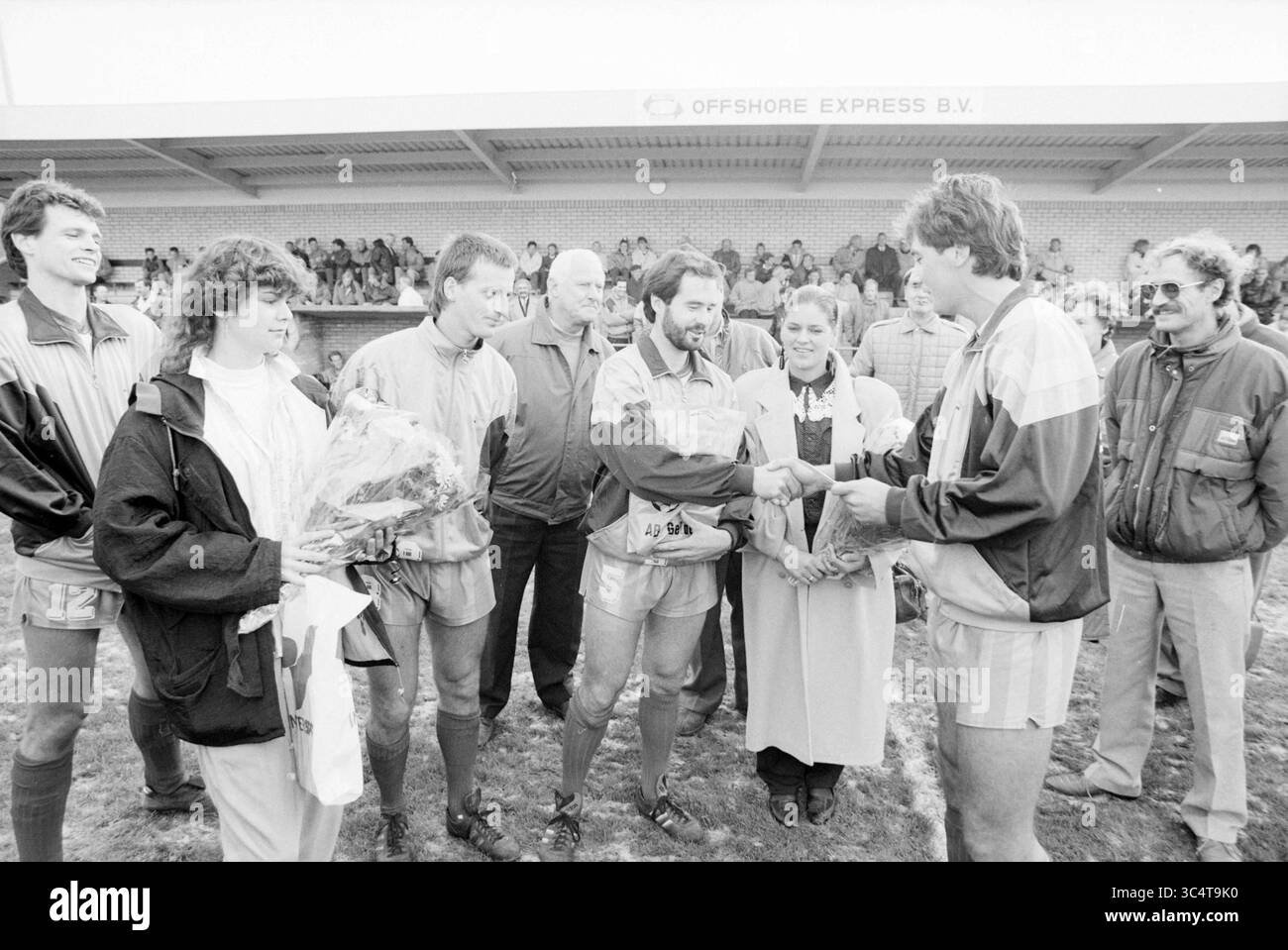 Wijk aan Zee, Football People and Trainer, Wijk aan Zee, 05-11-1989 Whizgle News, Dutch Desk, Niederlande, 1950-2000 Eine Gruppe von Athleten steht auf einem Sportplatz und interagiert mit einer Frau, die Blumen präsentiert. Die Szene fängt einen Moment der Feier ein, mit Lächeln und Gesten der Kameradschaft unter den Einzelpersonen, während die Zuschauer im Hintergrund zuschauen. Die Atmosphäre deutet auf eine Veranstaltung nach dem Spiel oder eine Preisverleihung hin. Stockfoto
