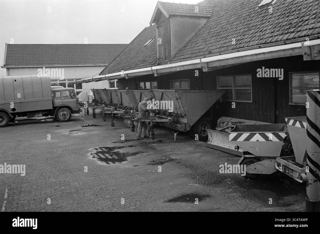 Reparatur von Schneeschaufeln in der Werkstatt der Gemeinde Velsen und Containern für die Salzstreuung., Velsen Whizgle News, Dutch Desk, Niederlande, 1950-2000 Eine Reihe von Industriemaschinen befindet sich neben einem Gebäude, bei dem eine einzelne Inspektion oder Arbeit an der Ausrüstung durchgeführt wird. In der Nähe steht ein altes Fahrzeug, und der Boden zeigt Anzeichen von neuem Regen mit Pfützen, die die Umgebung reflektieren. Stockfoto