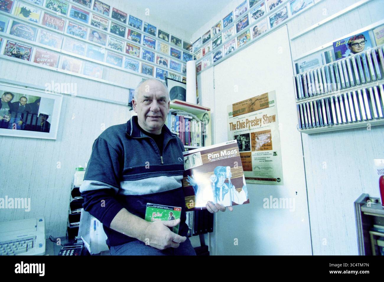 Hans Smit, Haarlem, Niederlande, 27-12-2000 Whizgle News, Dutch Desk, Niederlande, 1950-2000 Ein Mann sitzt in einem gemütlichen Raum voller Musik- und Erinnerungsstücke und hält eine Schallplatte mit dem Titel „Pint Moss“. Die Wände sind mit verschiedenen Musikplakaten und gerahmten Platten geschmückt, die eine tiefe Leidenschaft für Musik widerspiegeln. Stockfoto Hans Smit, Haarlem, Niederlande, 27-12-2000 Whizgle News, Dutch Desk, Niederlande, 1950-2000 Ein Mann sitzt in einem gemütlichen Raum voller Musik- und Erinnerungsstücke und hält eine Schallplatte mit dem Titel „Pint Moss“. Die Wände sind mit verschiedenen Musikplakaten und gerahmten Platten geschmückt, die eine tiefe Leidenschaft für Musik widerspiegeln. Stockfoto