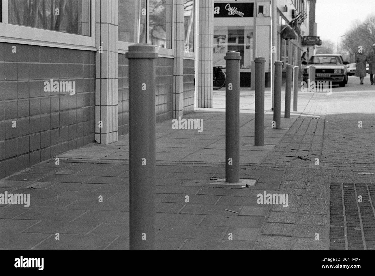 Straße mit Pfosten und Fußgängern plus Hund, 00-03-1990 Whizgle News, Dutch Desk, Niederlande, 1950-2000 Eine Reihe aufrechter Metallpfoller säumt einen Bürgersteig und steht im Kontrast zur urbanen Kulisse einer Straße, an der ein Auto vorbeifährt. Die Szene fängt einen Moment des alltäglichen Stadtlebens ein, eingerahmt von den Schaufensterfronten und der fernen Bewegung von Fußgängern. Stockfoto
