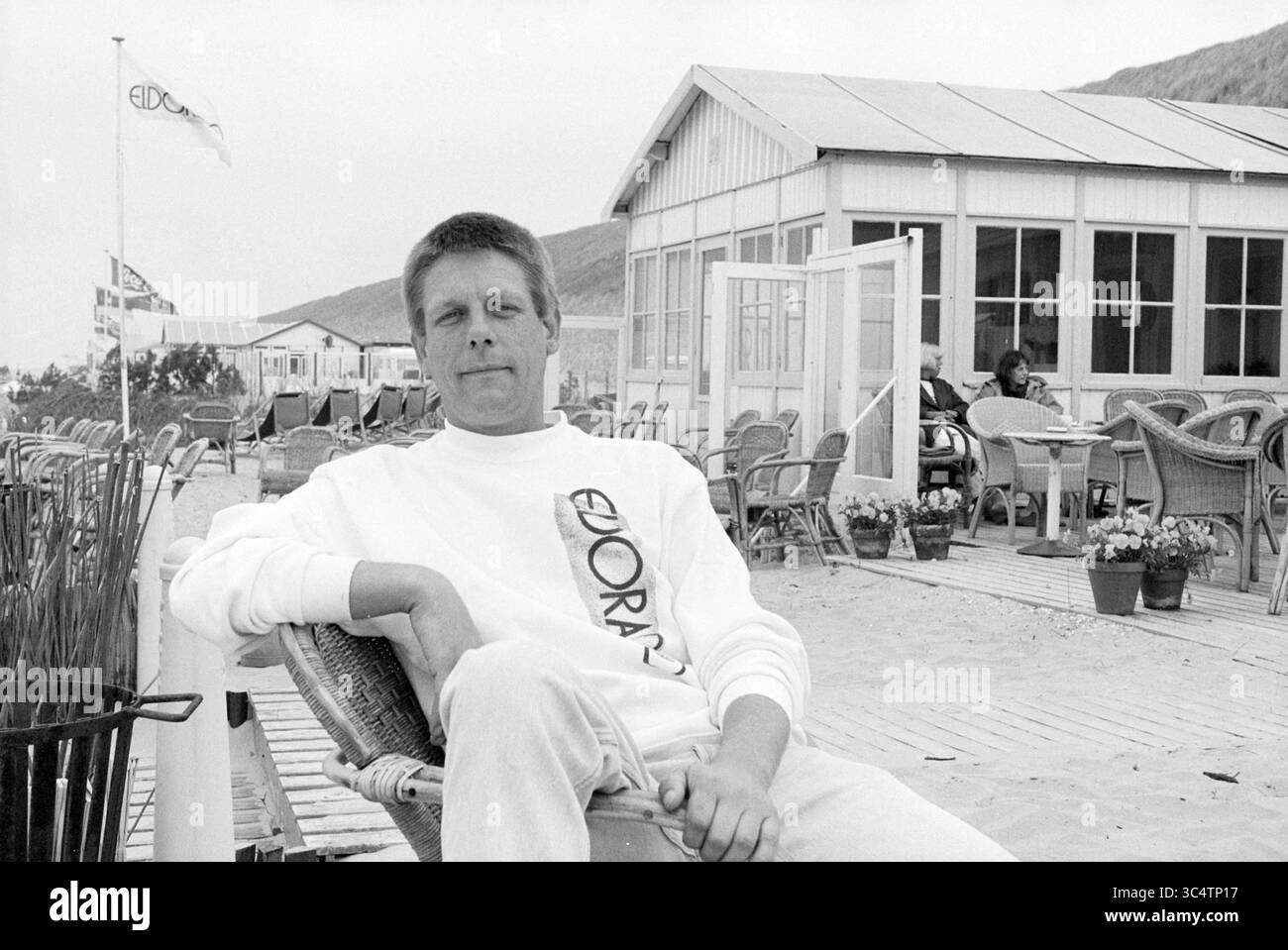 Jan Wanders, Z'voort, Zandvoort, 05.05.1995 Whizgle News, Dutch Desk, 31 Niederlande, 1950-2000 Ein entspannter Mann sitzt bequem in einem Sessel an einer Strandpromenade und trägt ein lässiges Sweatshirt. Hinter ihm ist ein bezauberndes Gebäude am Meer zu sehen, zusammen mit Menschen, die den Sitzbereich im Freien genießen und dekorative Pflanzen am Gehweg säumen. Stockfoto