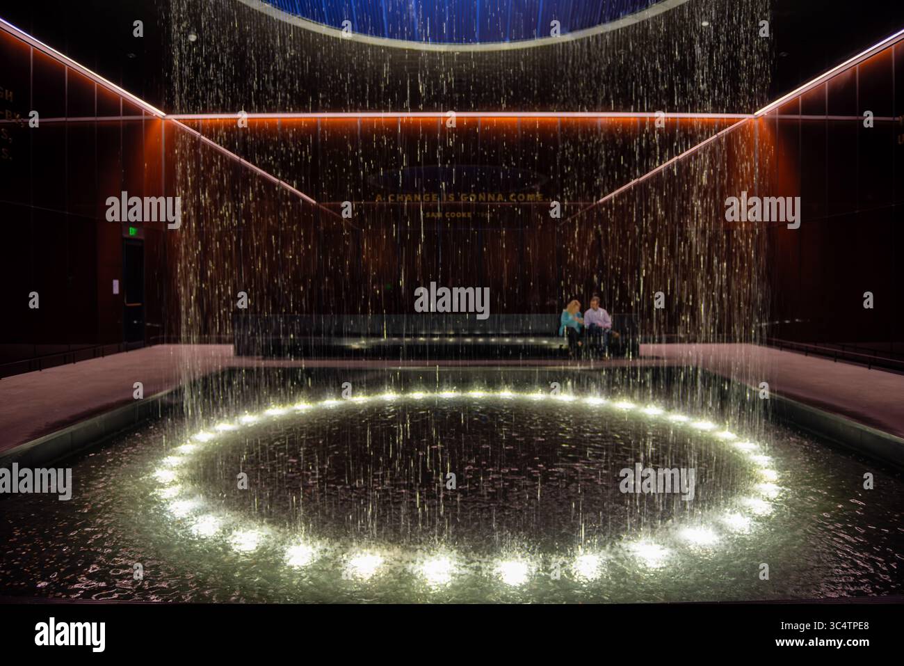 National Museum of African American History and Culture Contemplative Court Washington DC // WASHINGTON DC — der Contemplative Court im National Museum of African American History and Culture verfügt über einen zylindrischen Brunnen, in dem Wasser von einem runden Oberlicht in ein reflektierendes Becken unter sich fällt. Der Hof ist ein meditativer Übergangsraum und liegt zwischen den Geschichtsgalerien des Museums und den Ausstellungen zu kulturellen Feierlichkeiten. Er bietet Besuchern einen Moment der emotionalen Verarbeitung und Reflexion. Der Raum wird von markanten, karamellfarbenen Kupfer-Bronze-Glaswänden von Bendheim umschlossen Stockfoto