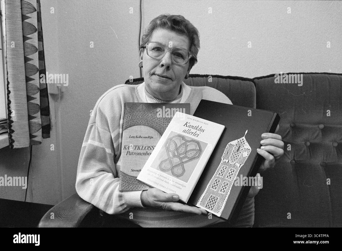 Mrs. Kolbe Buch Spulenspitze, Bücher und Buchhändler, 14-12-1989 Whizgle News, Dutch Desk, Niederlande, 1950-2000 Eine Frau sitzt auf einer Couch, hält zwei Bücher über traditionelles Handwerk, mit einem stolzen Lächeln. Ein Buch scheint sich auf Techniken der Schnürung zu konzentrieren, während das andere verschiedene Muster hervorhebt. Dekorative Elemente, darunter ein Lesezeichen aus Spitze, betonen ihre Darstellung. Stockfoto