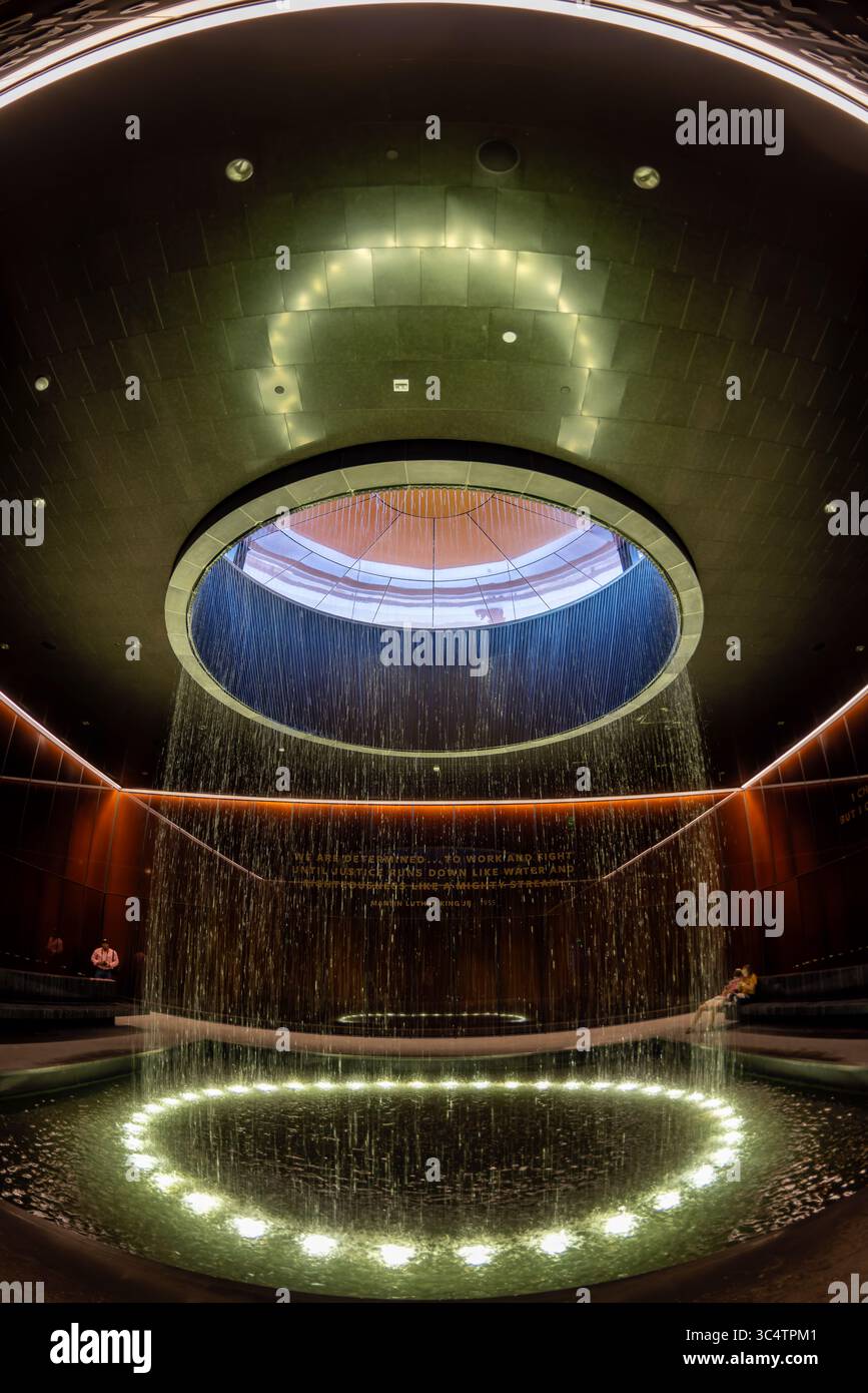 National Museum of African American History Contemplative Court Washington DC // WASHINGTON DC — der Contemplative Court im Smithsonian National Museum of African American History and Culture verfügt über einen zylindrischen Brunnen, der Wasser durch einen oculus mit Himmelslicht in einen kreisförmigen Pool unter sich gießt. Der Raum ist als Pausepunkt für Besucher konzipiert, die zwischen den emotional intensiven Geschichtsgalerien des Museums und seinen kulturellen Festausstellungen wechseln. Der Hof ist von kundenspezifischen, karamellfarbenen Kupfer-Bronze-Verbundglaswänden umgeben, die Bendheim in Zusammenarbeit mit der Architektur geschaffen hat Stockfoto