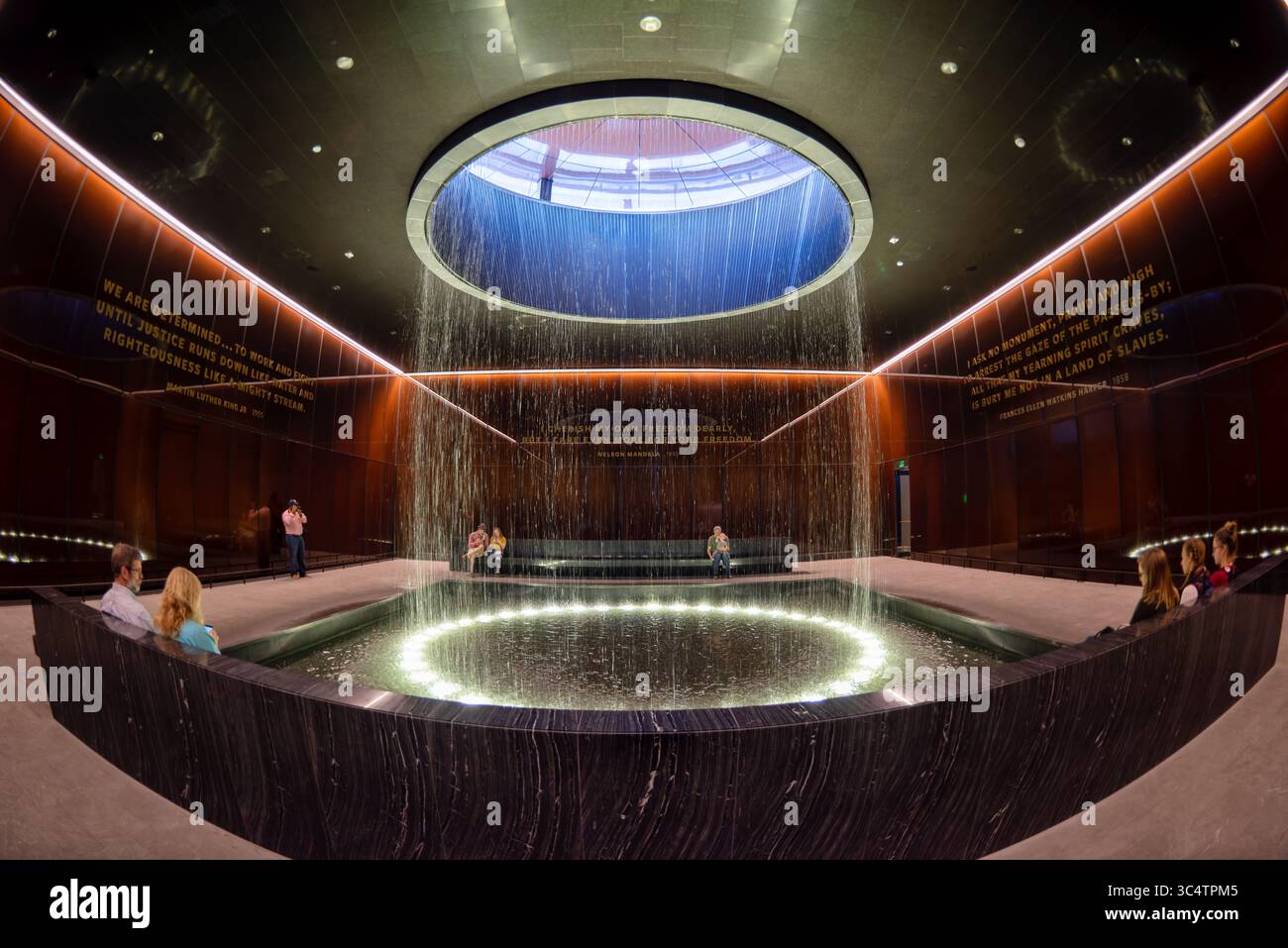 National Museum of African American History Contemplative Court Washington DC // WASHINGTON DC — der Contemplative Court im Smithsonian National Museum of African American History and Culture verfügt über einen zylindrischen Brunnen, der Wasser durch einen oculus mit Himmelslicht in einen kreisförmigen Pool unter sich gießt. Der Raum ist als Pausepunkt für Besucher konzipiert, die zwischen den emotional intensiven Geschichtsgalerien des Museums und seinen kulturellen Festausstellungen wechseln. Der Hof ist von kundenspezifischen, karamellfarbenen Kupfer-Bronze-Verbundglaswänden umgeben, die Bendheim in Zusammenarbeit mit der Architektur geschaffen hat Stockfoto