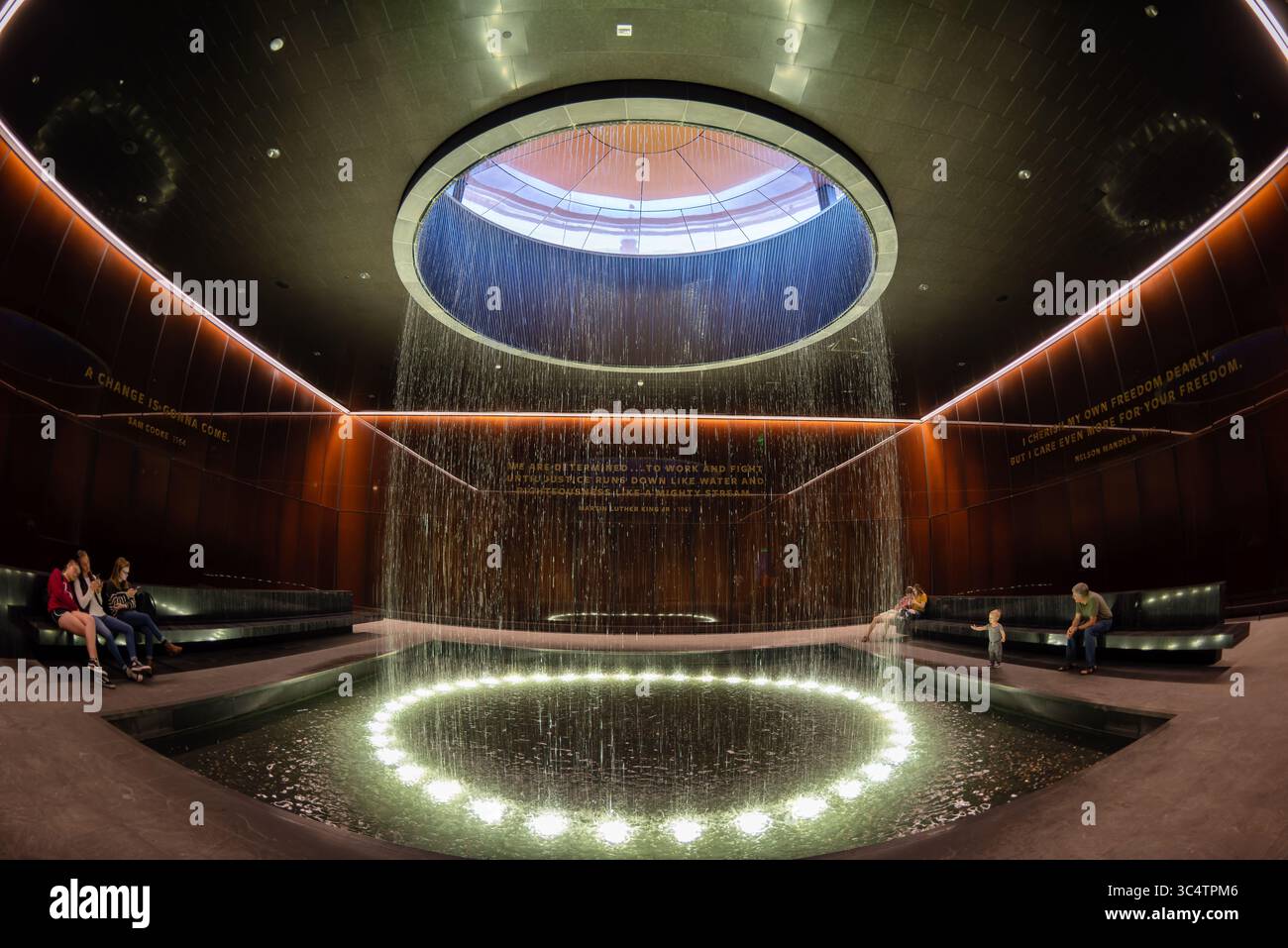 National Museum of African American History Contemplative Court Washington DC // WASHINGTON DC — der Contemplative Court im Smithsonian National Museum of African American History and Culture verfügt über einen zylindrischen Brunnen, der Wasser durch einen oculus mit Himmelslicht in einen kreisförmigen Pool unter sich gießt. Der Raum ist als Pausepunkt für Besucher konzipiert, die zwischen den emotional intensiven Geschichtsgalerien des Museums und seinen kulturellen Festausstellungen wechseln. Der Hof ist von kundenspezifischen, karamellfarbenen Kupfer-Bronze-Verbundglaswänden umgeben, die Bendheim in Zusammenarbeit mit der Architektur geschaffen hat Stockfoto