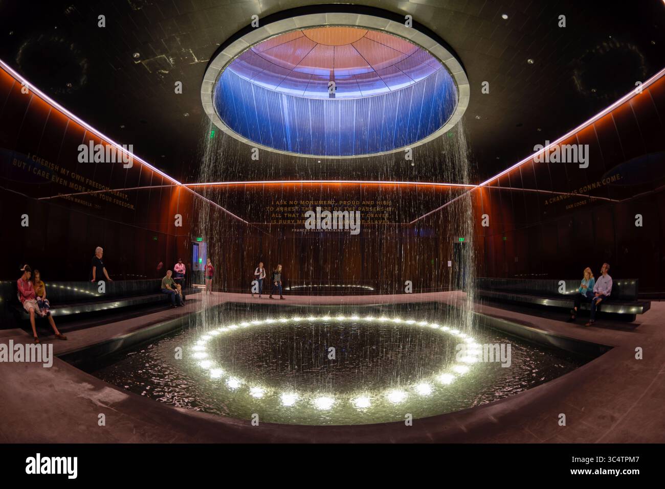 National Museum of African American History Contemplative Court Washington DC // WASHINGTON DC — der Contemplative Court im Smithsonian National Museum of African American History and Culture verfügt über einen zylindrischen Brunnen, der Wasser durch einen oculus mit Himmelslicht in einen kreisförmigen Pool unter sich gießt. Der Raum ist als Pausepunkt für Besucher konzipiert, die zwischen den emotional intensiven Geschichtsgalerien des Museums und seinen kulturellen Festausstellungen wechseln. Der Hof ist von kundenspezifischen, karamellfarbenen Kupfer-Bronze-Verbundglaswänden umgeben, die Bendheim in Zusammenarbeit mit der Architektur geschaffen hat Stockfoto