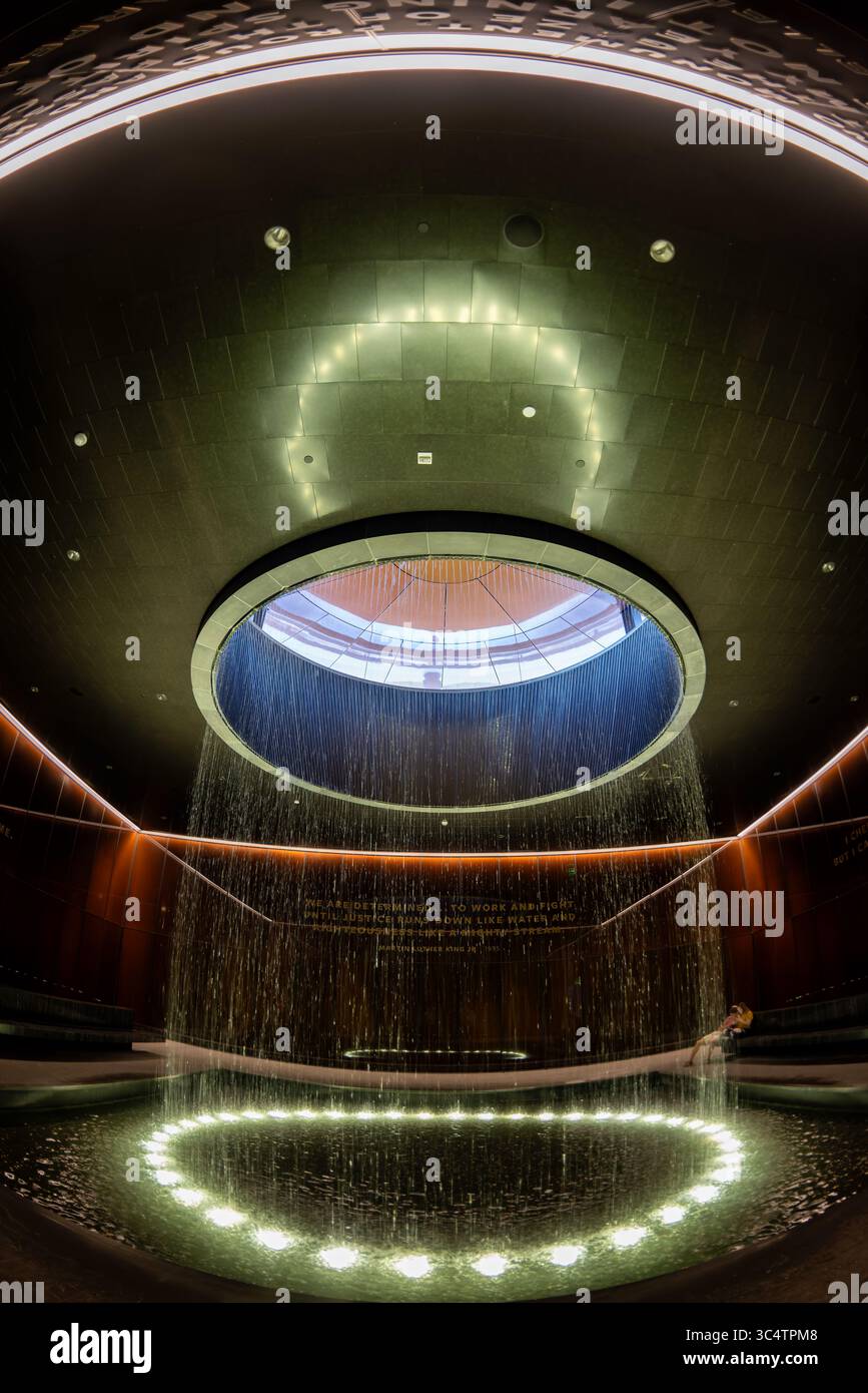 National Museum of African American History Contemplative Court Fountain Washington DC // WASHINGTON DC — der Contemplative Court im Smithsonian National Museum of African American History and Culture verfügt über einen zylindrischen Brunnen, der Wasser durch einen oculus mit Himmelslicht in einen kreisförmigen Pool unter sich gießt. Der Raum ist als Pausepunkt für Besucher konzipiert, die zwischen den emotional intensiven Geschichtsgalerien des Museums und seinen kulturellen Festausstellungen wechseln. Der Hof ist von kundenspezifischen, karamellfarbenen Kupfer-Bronze-Verbundglaswänden umgeben, die Bendheim in Zusammenarbeit mit der AR geschaffen hat Stockfoto