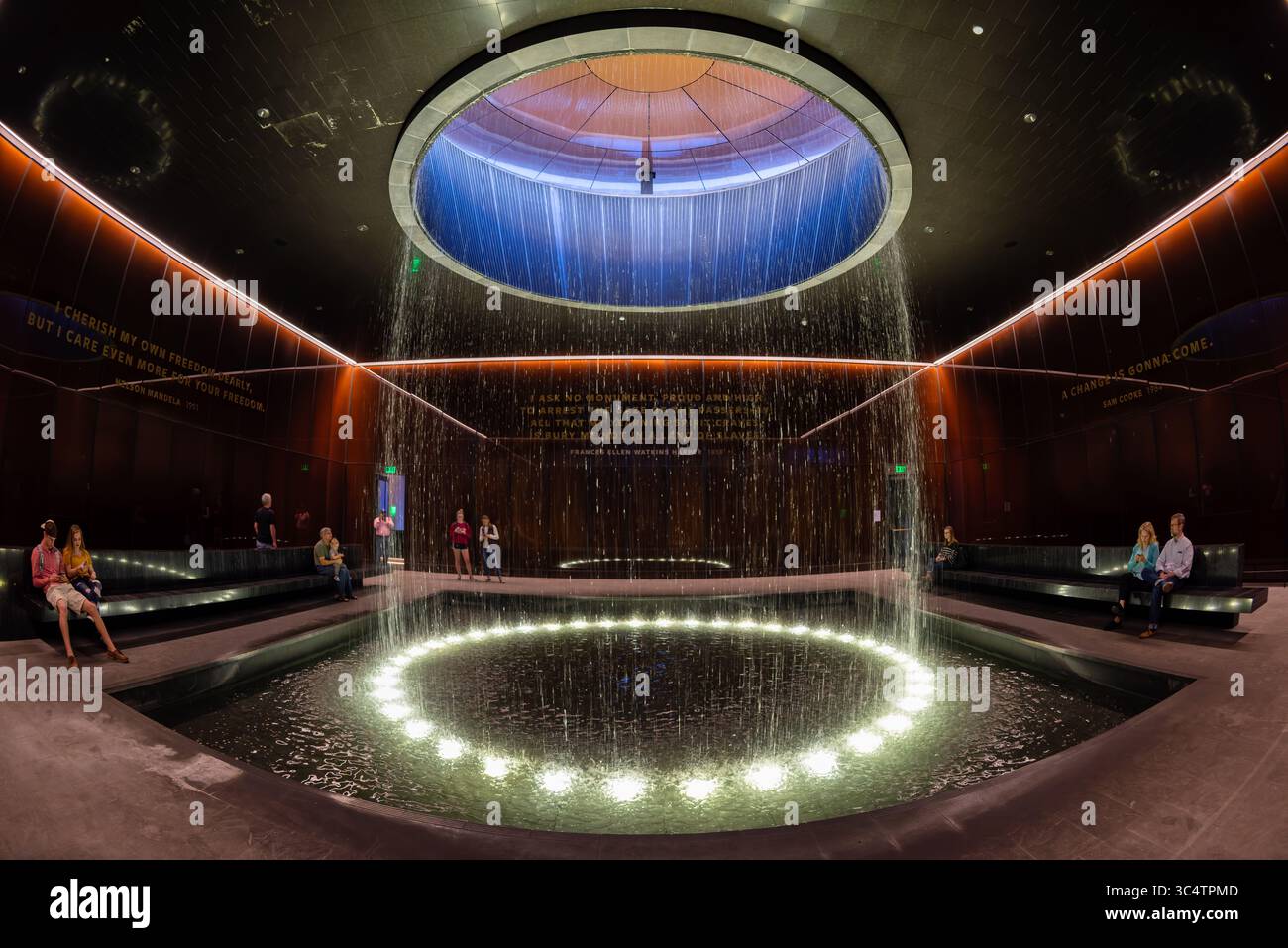 National Museum of African American History Contemplative Court Washington DC // WASHINGTON DC — der Contemplative Court im Smithsonian National Museum of African American History and Culture verfügt über einen zylindrischen Brunnen, der Wasser durch einen oculus mit Himmelslicht in einen kreisförmigen Pool unter sich gießt. Der Raum ist als Pausepunkt für Besucher konzipiert, die zwischen den emotional intensiven Geschichtsgalerien des Museums und seinen kulturellen Festausstellungen wechseln. Der Hof ist von kundenspezifischen, karamellfarbenen Kupfer-Bronze-Verbundglaswänden umgeben, die Bendheim in Zusammenarbeit mit der Architektur geschaffen hat Stockfoto
