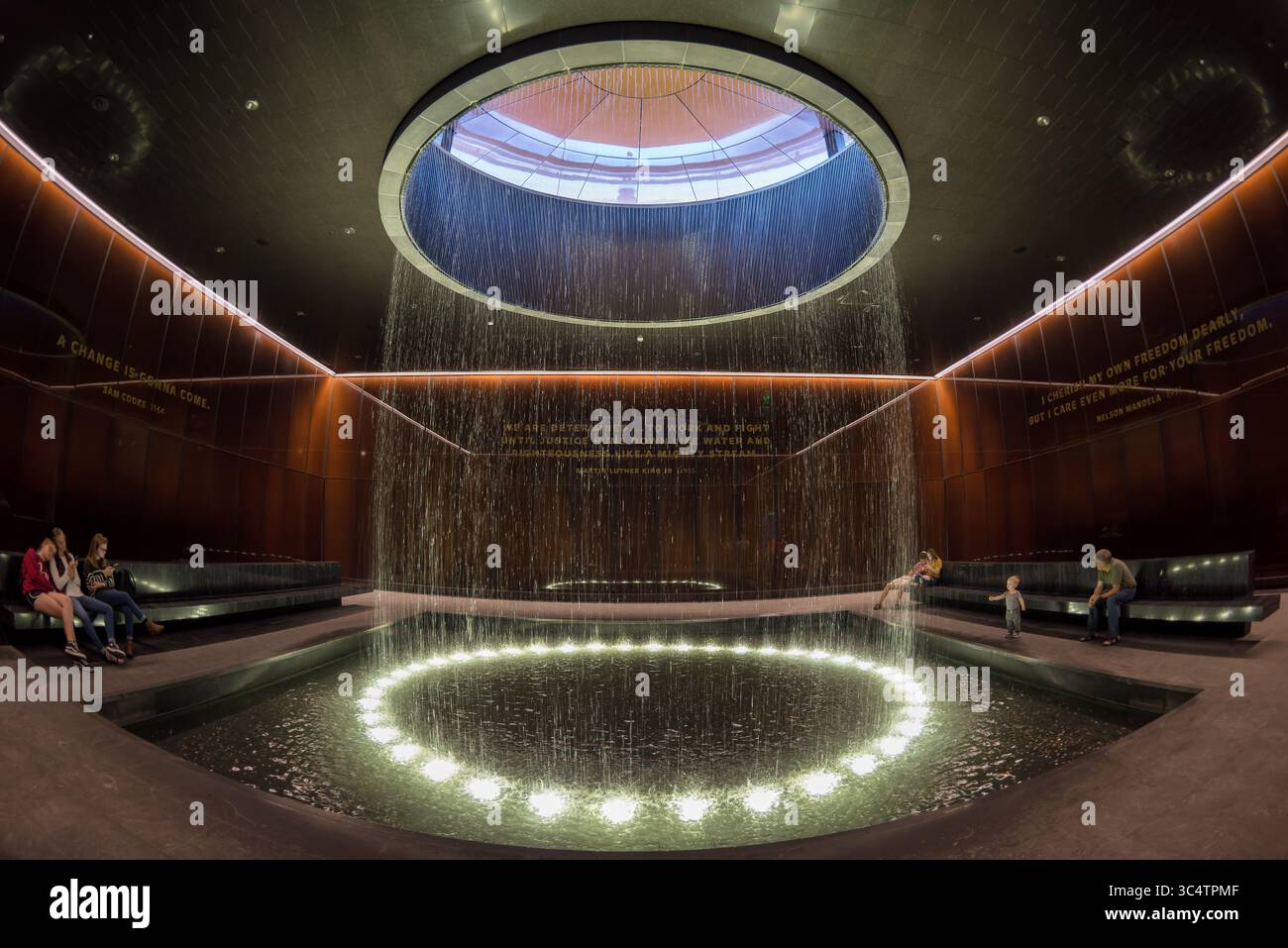 National Museum of African American History Contemplative Court Washington DC // WASHINGTON DC — der Contemplative Court im National Museum of African American History and Culture verfügt über einen zylindrischen Brunnen, in dem Wasser von einem runden Oberlicht in ein reflektierendes Becken fällt. Der Hof ist ein meditativer Übergangsraum und liegt zwischen den Geschichtsgalerien des Museums und den Ausstellungen zu kulturellen Feierlichkeiten. Er bietet Besuchern einen Moment der emotionalen Verarbeitung und Reflexion. Der Raum wird von markanten, karamellfarbenen Kupfer-Bronze-Glaswänden umschlossen, die Bendheim in Kollabor geschaffen hat Stockfoto