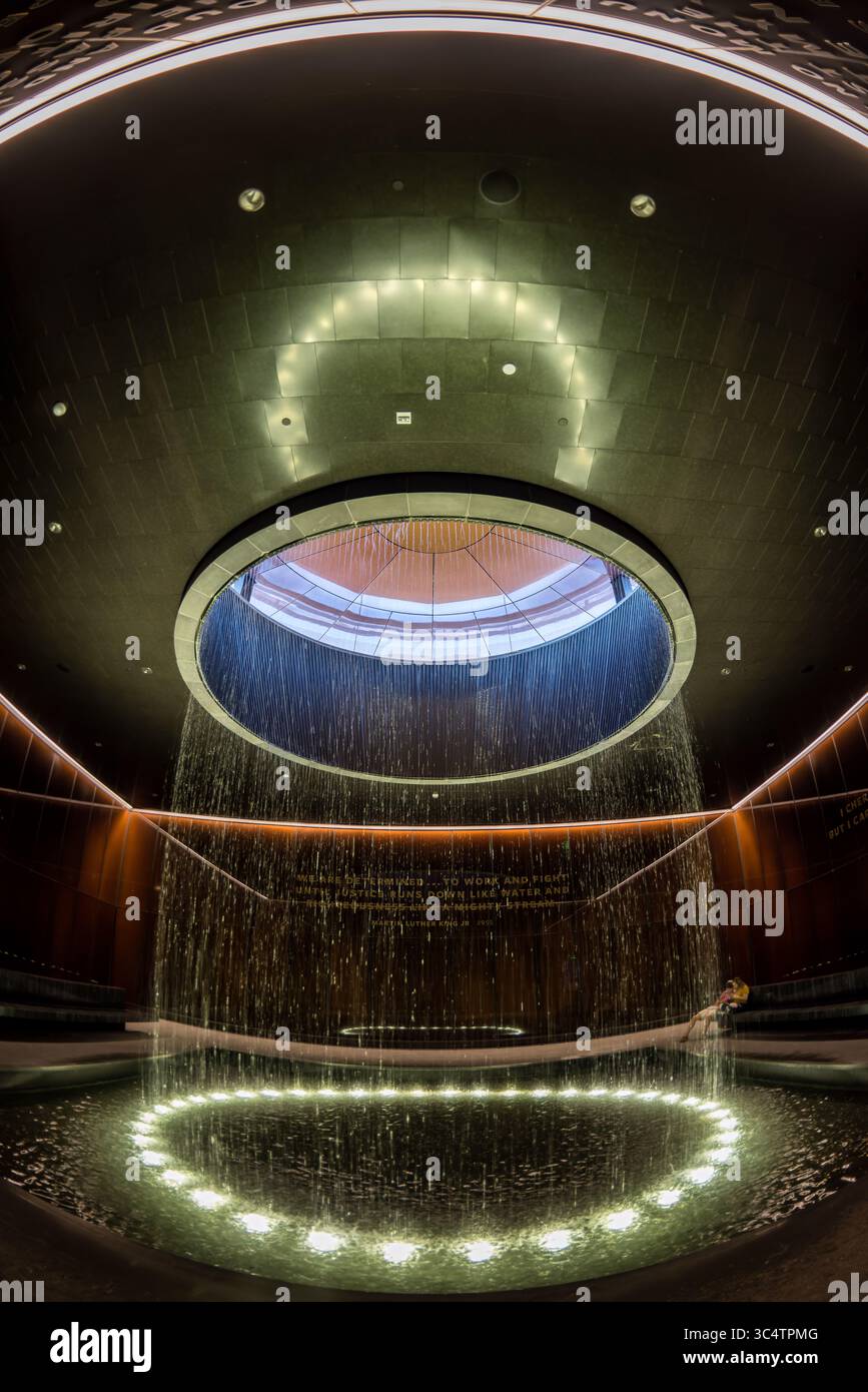 National Museum of African American History Contemplative Court Washington DC // WASHINGTON DC — der Contemplative Court im National Museum of African American History and Culture verfügt über einen zylindrischen Brunnen, in dem Wasser von einem runden Oberlicht in ein reflektierendes Becken fällt. Der Hof ist ein meditativer Übergangsraum und liegt zwischen den Geschichtsgalerien des Museums und den Ausstellungen zu kulturellen Feierlichkeiten. Er bietet Besuchern einen Moment der emotionalen Verarbeitung und Reflexion. Der Raum wird von markanten, karamellfarbenen Kupfer-Bronze-Glaswänden umschlossen, die Bendheim in Kollabor geschaffen hat Stockfoto