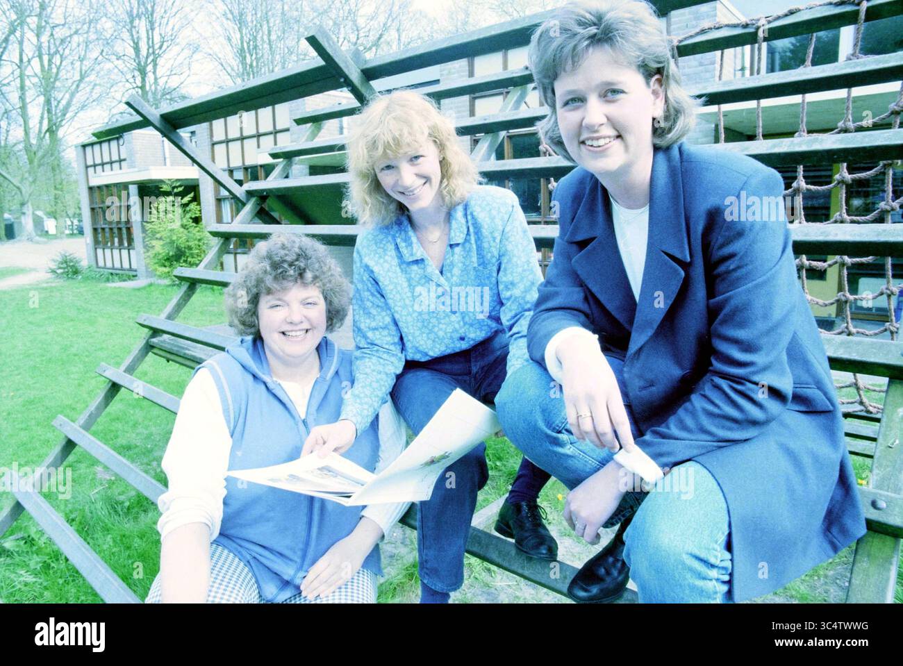 Drei Damen mit Buch MKD Margriet [medizinische Kindertagesstätte], 20-04-1998 Whizgle News, Dutch Desk, Niederlande, 1950-2000 drei Frauen versammeln sich draußen, lächelnd und verwickelt sich in Gespräche. Eine Frau sitzt auf dem Boden, während die anderen neben ihr knien und Dokumente halten. Sie sind von einer natürlichen Umgebung mit Grün und einer architektonischen Kulisse umgeben. Jede Frau hat eine eigene Frisur und trägt legere Kleidung, die eine freundliche und gemeinschaftliche Atmosphäre vermittelt. Stockfoto