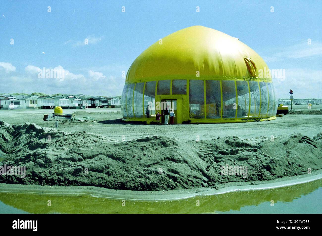 Theater am Meer, IJmuiden, Niederlande, 20-05-2003 Whizgle News, Dutch Desk, Niederlande, 1950-2000 Ein großer, kuppelförmiger Bau mit hellgelber Fassade steht auf sandigem Gelände, umgeben von kleinen, dezenten Gebäuden. Zwei Personen sind in der Nähe des Eingangs zu sehen, während die Reflexionen in einem nahe gelegenen Wasserbecken schimmern. Die Landschaft weist sanfte Hügel im Hintergrund auf, die eine einzigartige und unkonventionelle Umgebung darstellen. Stockfoto