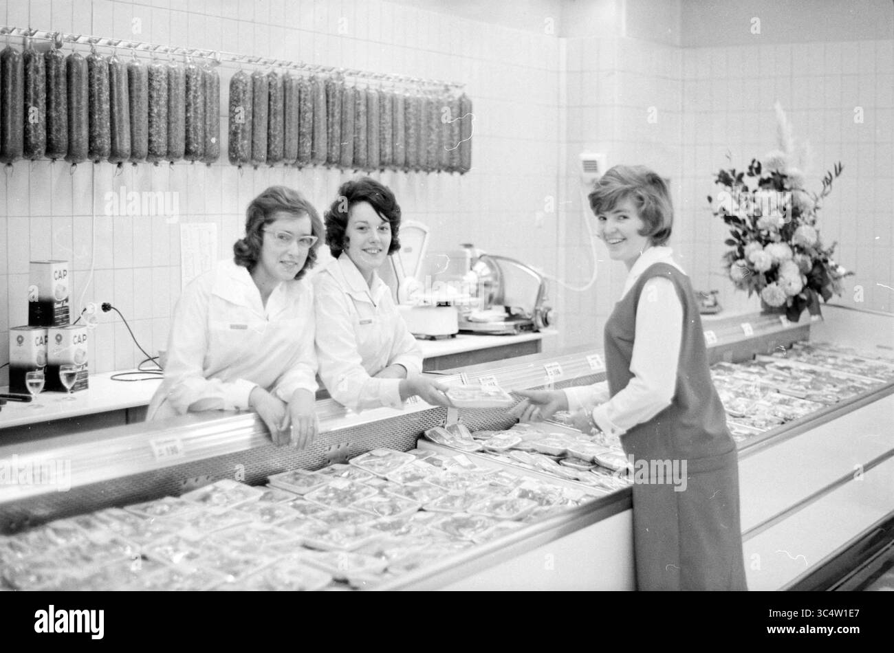 Metzgerabteilung Supermarkt, 14-11-1962 Whizgle News, Dutch Desk, Niederlande, 1950-2000 drei Frauen arbeiten hinter einer Delikatessentheke, die mit verschiedenen Fleischprodukten und verpackten Waren gefüllt ist. Sie lächeln und interagieren mit einem Kunden, der sich über den Tresen beugt, um eine Auswahl zu treffen. Im Hintergrund hängen Salamis, und ein dekoratives Blumendesign verleiht der Szene einen Hauch Eleganz. Stockfoto