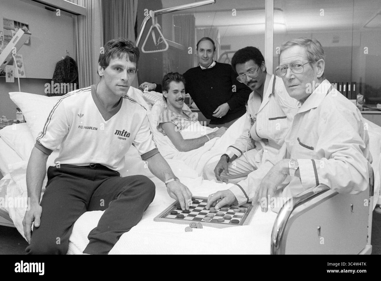 Martin Haar in Maria Foundation, Football Haarlem, 08-12-1987 Whizgle News, Dutch Desk, Niederlande, 1950-2000 Eine Gruppe von fünf Männern versammelt sich um ein Krankenhausbett, wobei ein Mann auf dem Bett liegt, während vier andere in der Nähe sitzen. Sie sind in einem Spiel von Schachspielern verwickelt und zeigen einen Moment der Kameradschaft und der Freizeit trotz der Krankenhaussituation. Die Gesichtsausdrücke spiegeln eine Mischung aus Konzentration, Unterhaltung und Unterstützung wider. Stockfoto