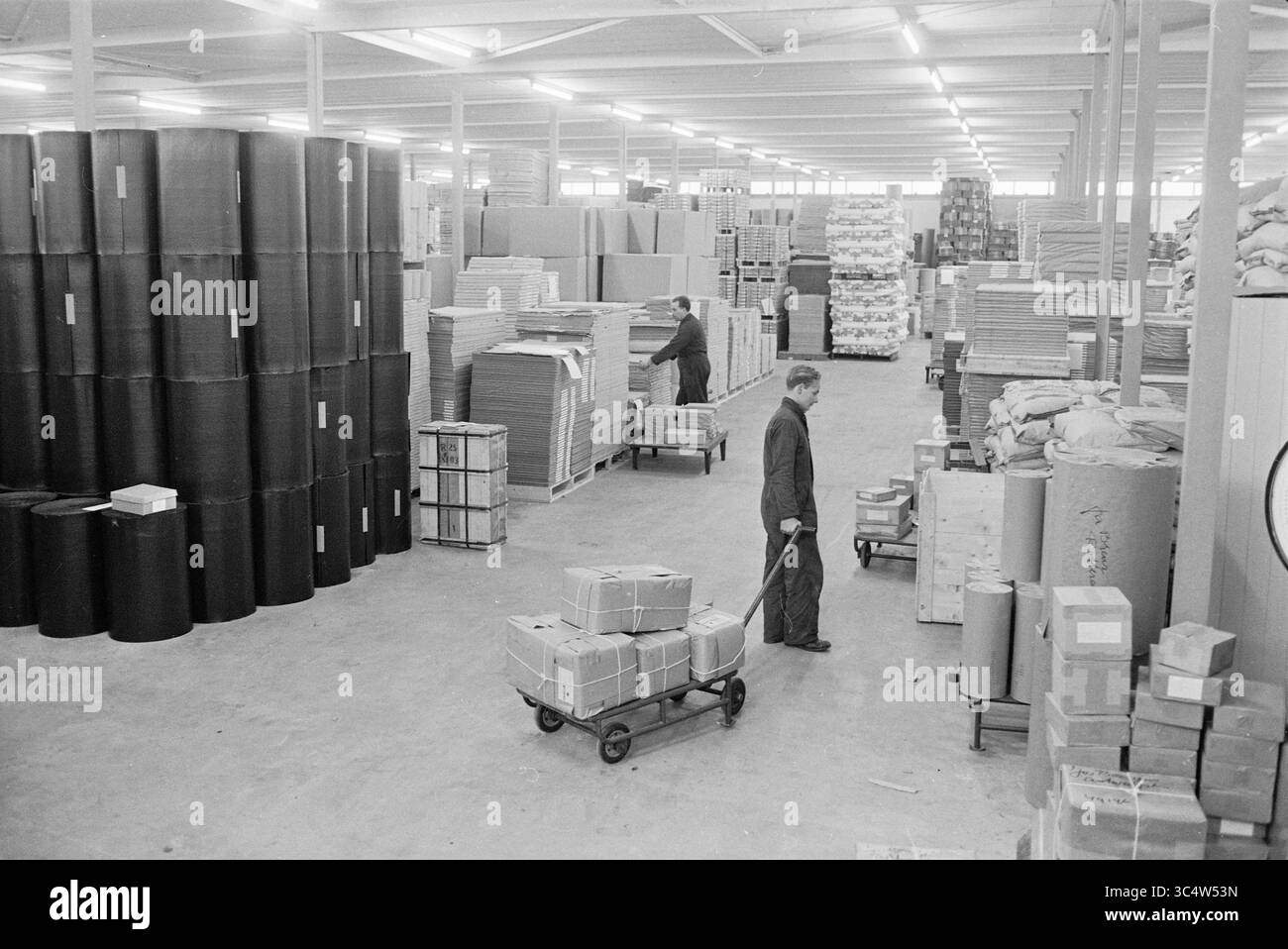FA.Ruys Verpackungsmaterial kleine Houtweg Haarlem, Interieur neue und alte Fabrik, Fabriken, Fertigung, Werkstätten, Haarlem, Kleine Houtweg, Niederlande, 20-12-1962 Whizgle News, Dutch Desk, Niederlande, 1950-2000 Ein geräumiges Lager mit Stapeln verpackter Waren, einschließlich großer Rollen und Kisten. Arbeiter werden beim Manövrieren von Wagen beobachtet, die das Inventar zwischen den hoch aufragenden Materialstapeln organisieren. Die Szene spiegelt eine geschäftige und effiziente Speicherumgebung wider. Stockfoto