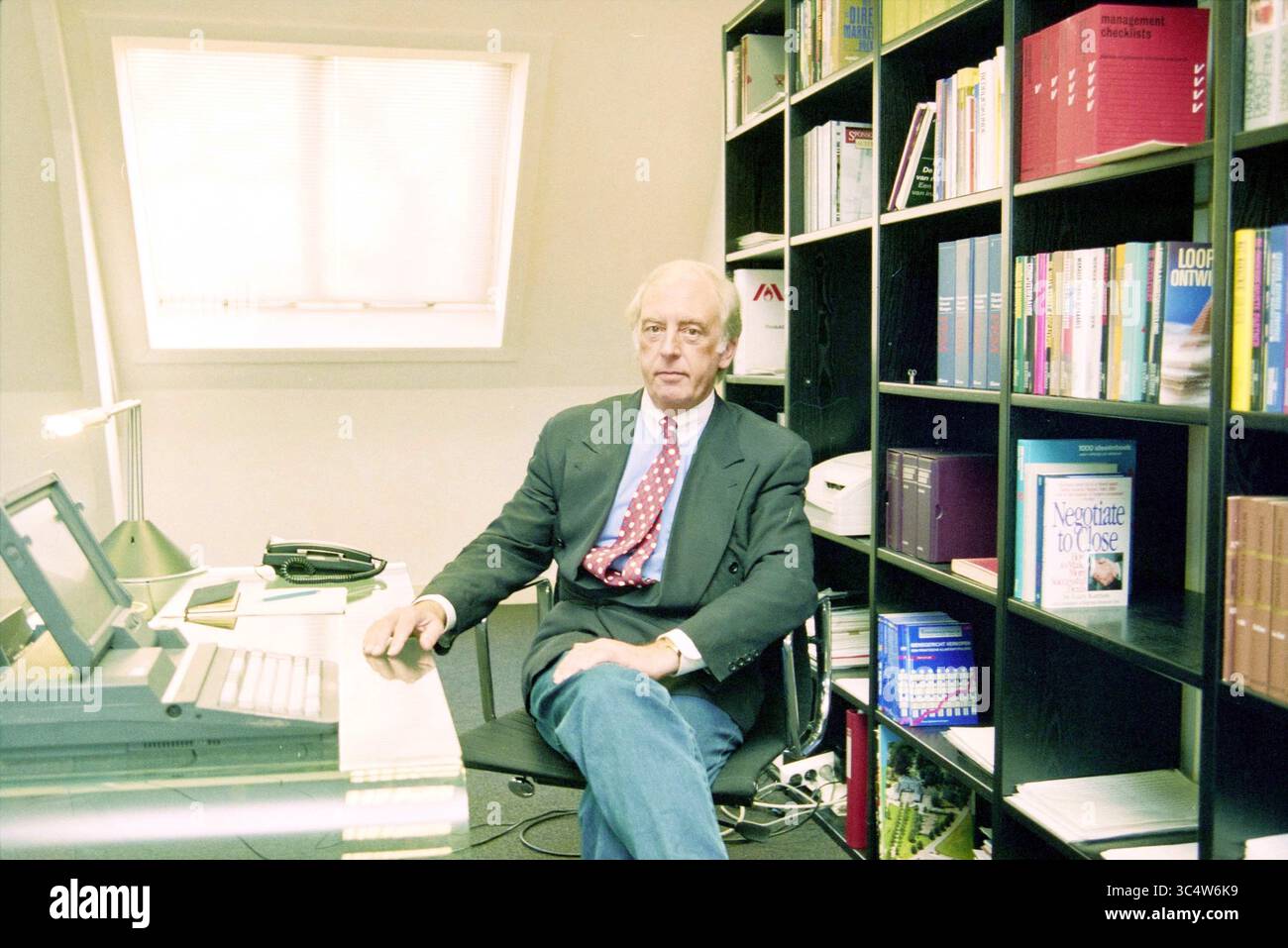 Jean Smith, 30-01-1996 Whizgle News, Dutch Desk, Niederlande, 1950-2000 Ein angesehener Mann sitzt an einem Schreibtisch in einem modernen Büro, umgeben von ordentlich angeordneten Bücherregalen mit verschiedenen Texten. Er trägt formelle Kleidung und strahlt einen Hauch von Professionalität und Selbstvertrauen aus. Der Arbeitsbereich verfügt über einen Computer, ein Telefon und ein gut organisiertes Layout, was eine produktive Umgebung suggeriert. Stockfoto