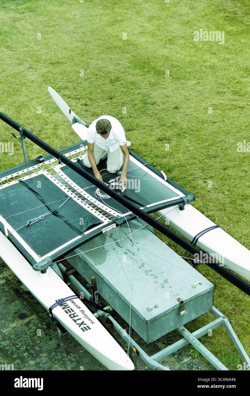 Thorin Sailmaker, Bloemendaal, 29-07-2003 Whizgle News, Dutch Desk, Niederlande, 1950-2000 Eine Person bereitet einen Katamaran für das Segeln vor und passt die Takelage auf dem Deck an, während sie von grasbewachsenem Gelände umgeben ist. Stockfoto