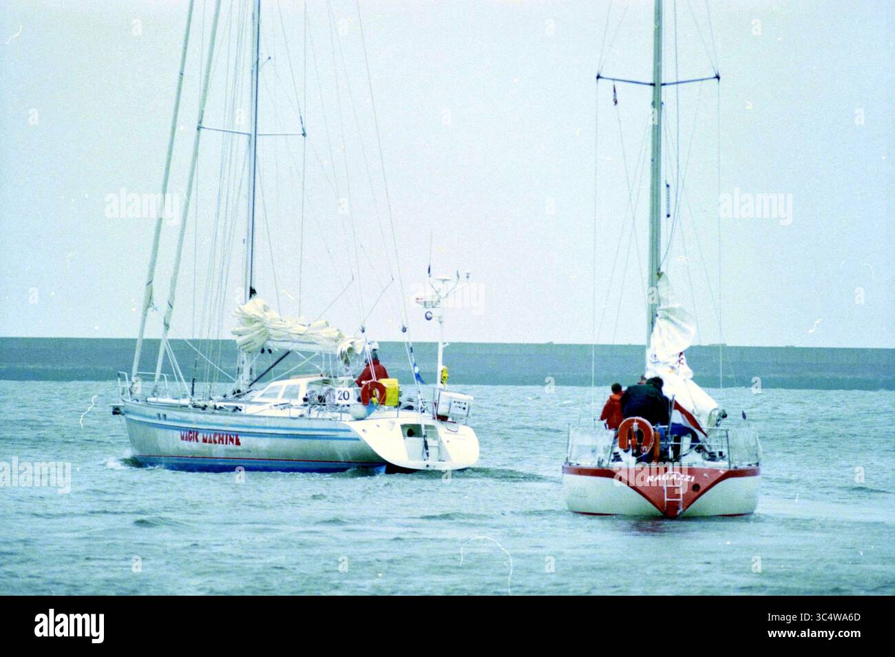 Start des Segelrennens in IJmuiden, IJmuiden, Niederlande, 12-07-1996 Whizgle News, Dutch Desk, Niederlande, 1950-2000 zwei Segelboote navigieren durch das Wasser, wobei ein Boot einen hellroten Rumpf und das andere den Namen „Mast Greeted“ trägt. Seeleute auf beiden Schiffen passen die Segel an, während sie über die Wellen gleiten, umgeben von einem ruhigen Meer. Ein entfernter Wellenbrecher ist sichtbar, der ein Gefühl der Zuflucht im offenen Wasser bietet. Stockfoto