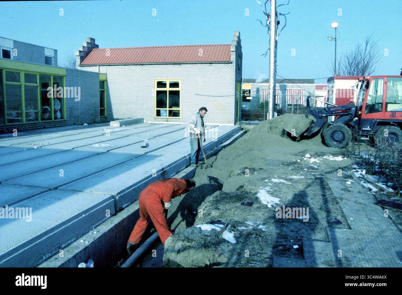 Bau von Schulräumen, Velserbroek, 11-02-1999 Whizgle News, Dutch Desk, Niederlande, 1950-2000 Arbeiter arbeiten im Baugewerbe, wobei einer neben einem Sandhaufen gräbt, während ein anderer in der Nähe schwere Maschinen bedient. Im Hintergrund steht ein Gebäude mit grün getönten Fenstern, das die Szene der aktiven Entwicklung abrundet. Stockfoto
