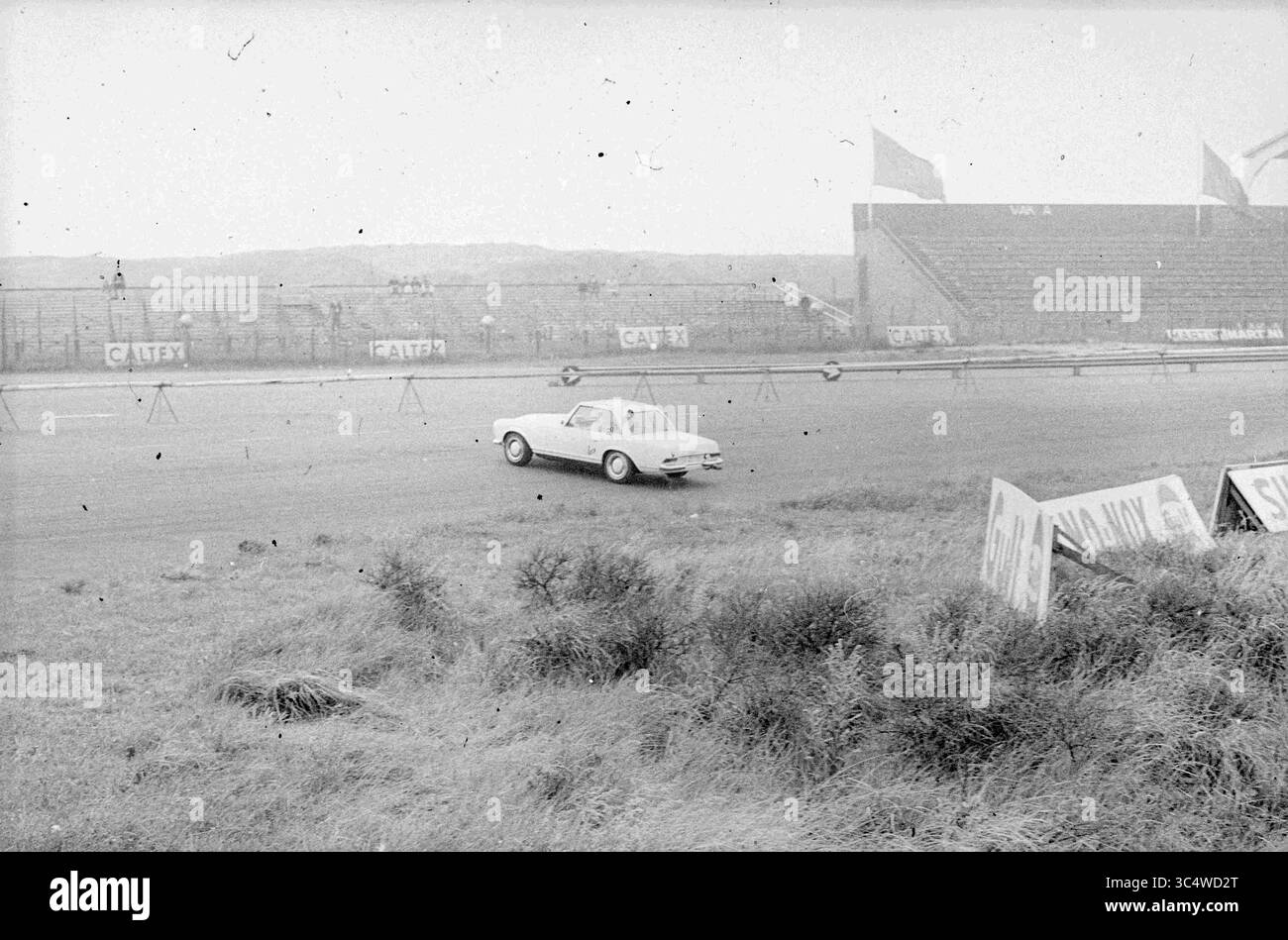 Autos auf der Strecke von Zandvoort, Zandvoort Whizgle News, Dutch Desk, Niederlande, 1950-2000 Ein einsames Auto fährt auf einer staubigen Strecke, umgeben von leeren Tribünen und bewachsenem Gras, was ein Gefühl der Nostalgie für den Oldtimer-Motorsport weckt. Stockfoto