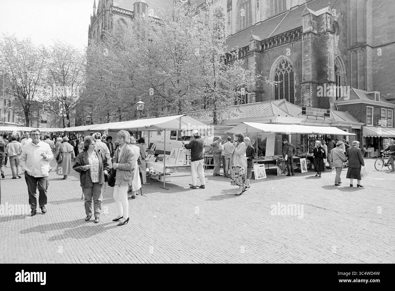 Kunstmarkt am Grote Markt, Grote Markt, Markt, Haarlem, Grote Markt, Niederlande, 11-05-1989 Whizgle News, Dutch Desk, Niederlande, 1950-2000 Eine geschäftige Marktszene mit zahlreichen Ständen mit Händlern und Käufern, die sich unter Bäumen mischen, vor dem Hintergrund eines beeindruckenden gotischen Gebäudes. Die Menschen führen Gespräche, durchsuchen Waren und genießen die lebhafte Atmosphäre des Marktes. Stockfoto
