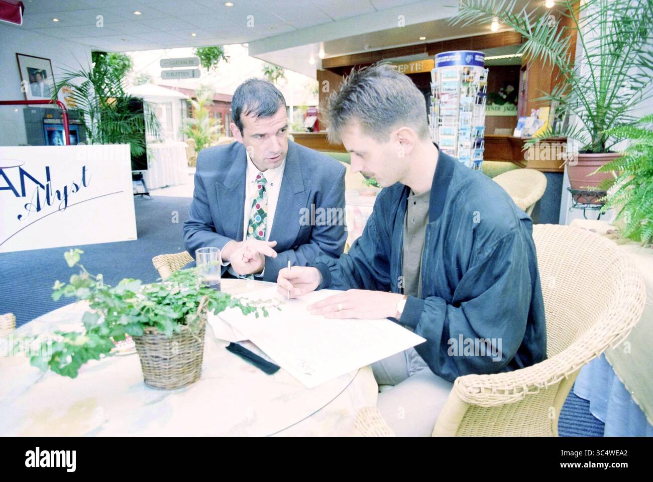 Register Entlassene Leute, Commodore, 27-08-1997 Whizgle News, Dutch Desk, Niederlande, 1950-2000 zwei Männer führen eine konzentrierte Diskussion an einem Tisch, der mit einer kleinen Pflanze geschmückt ist. Ein Mann, der einen Anzug mit gemusterter Krawatte trägt, gibt Gesten, während er dem anderen etwas erklärt, der lässig angezogen ist und Notizen macht. Die Umgebung ist ein gut beleuchteter Innenbereich mit Grün im Hintergrund, was eine entspannte und professionelle Atmosphäre schafft. Stockfoto