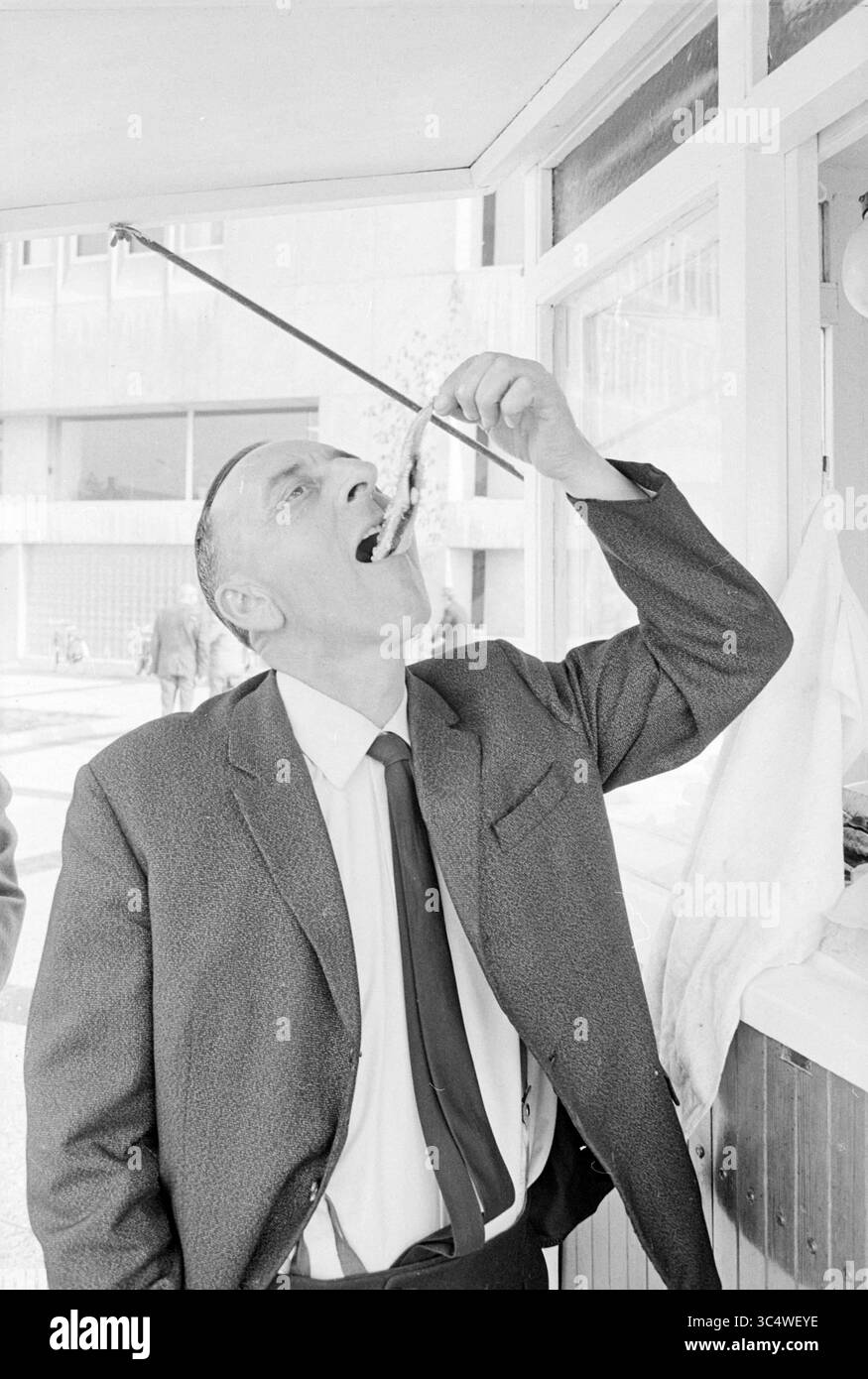 Fisch fressender Mann, Personen, 12-05-1969 Whizgle News, Dutch Desk, Niederlande, 1950-2000 Ein Mann in einem Anzug hebt mit einem langen Stock Essen in den Mund und zeigt einen amüsanten Moment des Essens. Er steht in der Nähe eines Essensfensters, mit Menschen und einem Gebäude im Hintergrund. Stockfoto