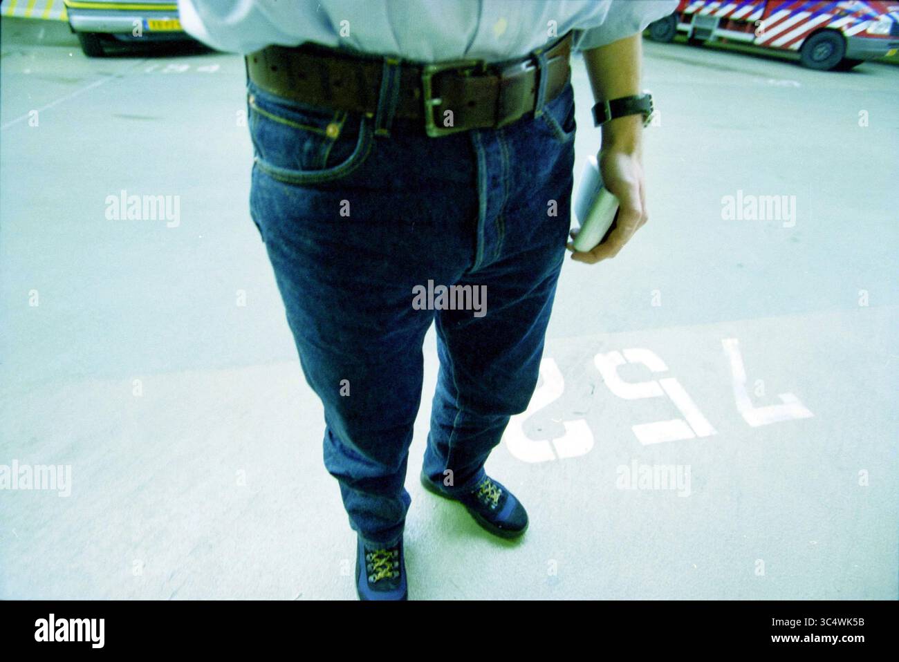 Mann in Jeans, 00-00-2001 Whizgle News, Dutch Desk, Niederlande, 1950-2000 Eine Person steht auf einem Parkplatz, trägt ein weißes Hemd in hochgeschnittenen blauen Jeans, gesichert mit einem braunen Gürtel. Ihre Füße sind in dunkle Schuhe gekleidet, und sie halten einen kleinen Gegenstand in der rechten Hand. Der Boden ist mit einer sichtbaren Zahl markiert. Stockfoto