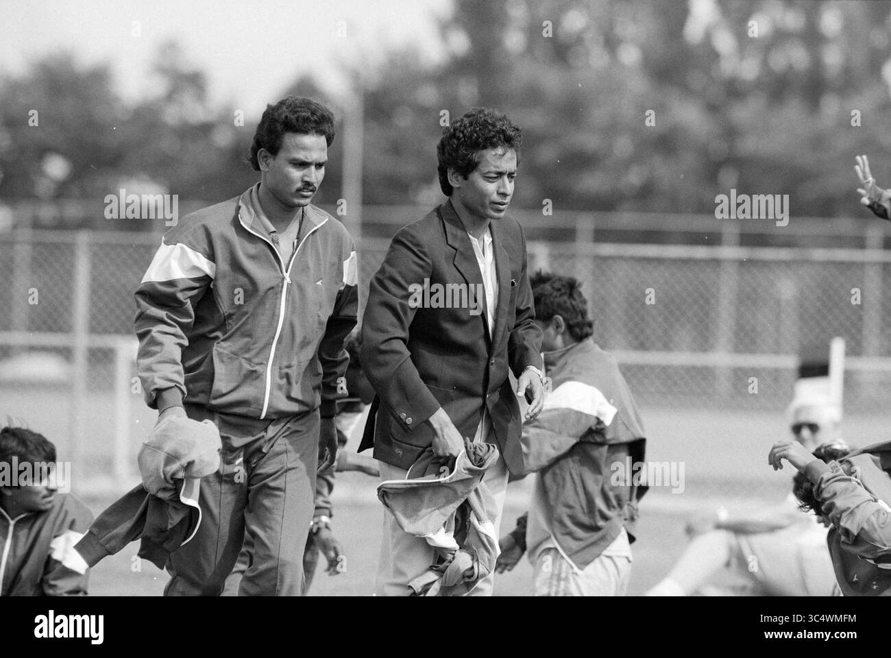 Hollandia - Indien + Regisseur + Trainer Indien, 08-08-1991 Whizgle News, Dutch Desk, Niederlande, 1950-2000 zwei Männer laufen über ein Sportfeld, einer in sportlicher Kleidung und der andere in einer formellen Jacke. Im Hintergrund ist eine Gruppe von Einzelpersonen an Aktivitäten beteiligt, die möglicherweise ein Spiel ausüben oder sich auf ein Spiel vorbereiten. Bäume und ein verschwommener Zaun bilden eine Kulisse, die auf eine lebhafte Atmosphäre im Sport hinweist. Stockfoto