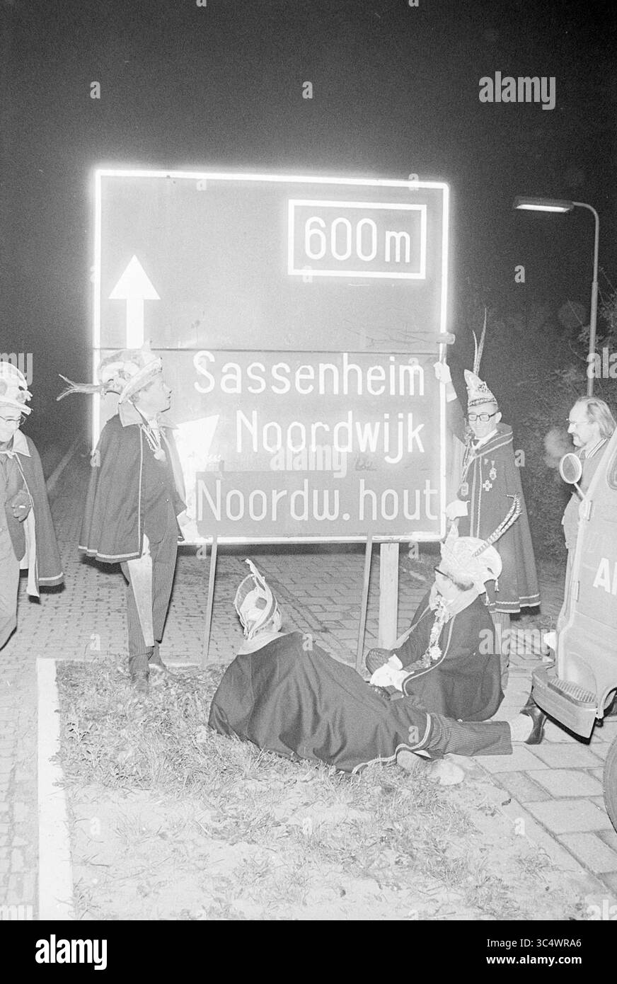 Anbringung von ANWB-Schildern für Prince Carnival, Carnival, 06-11-1969 Whizgle News, Dutch Desk, Niederlande, 1950-2000 Eine Gruppe von Menschen, die in festlichen Kostümen und Hüten gekleidet sind, versammelt sich um ein Straßenschild, das den Weg nach Sassenheim und Noordwijk anzeigt, während eine Person auf dem Boden sitzt. Die Szene spielt nachts, beleuchtet durch die Neonschilder oben. Stockfoto